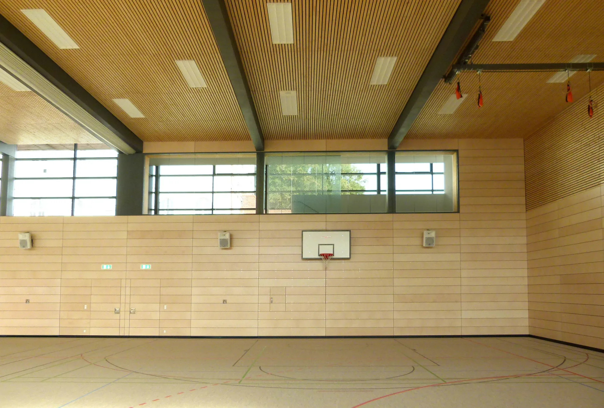 Blick in eine leere Turnhalle mit Holzdecken, großen Fenstern und einem Basketballkorb an der Wand.