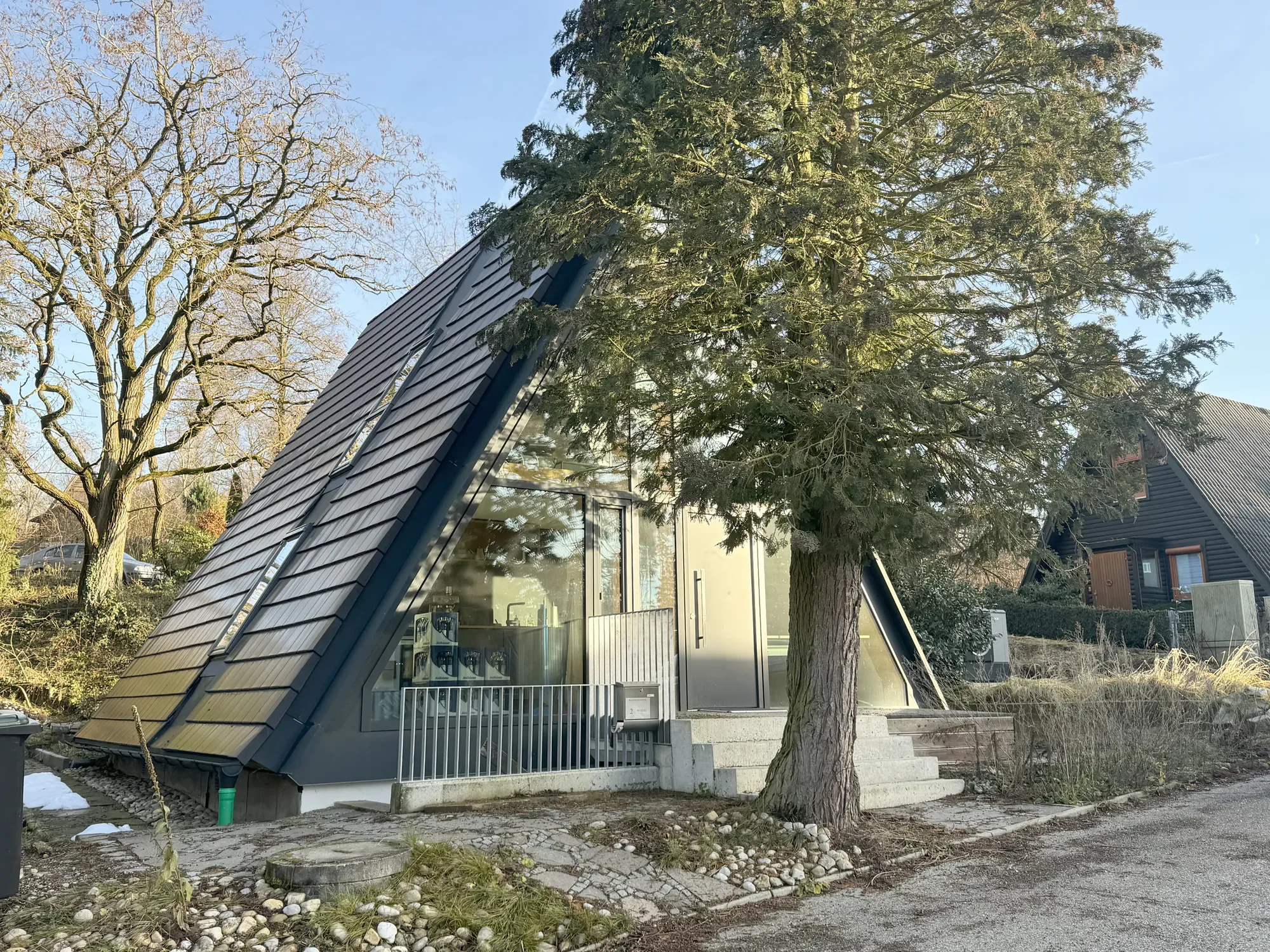 Modernes, schräg stehendes Haus mit dunklen Ziegeldach, große Fenster und Steinstufen, umgeben von Bäumen und einem klaren blauen Himmel.