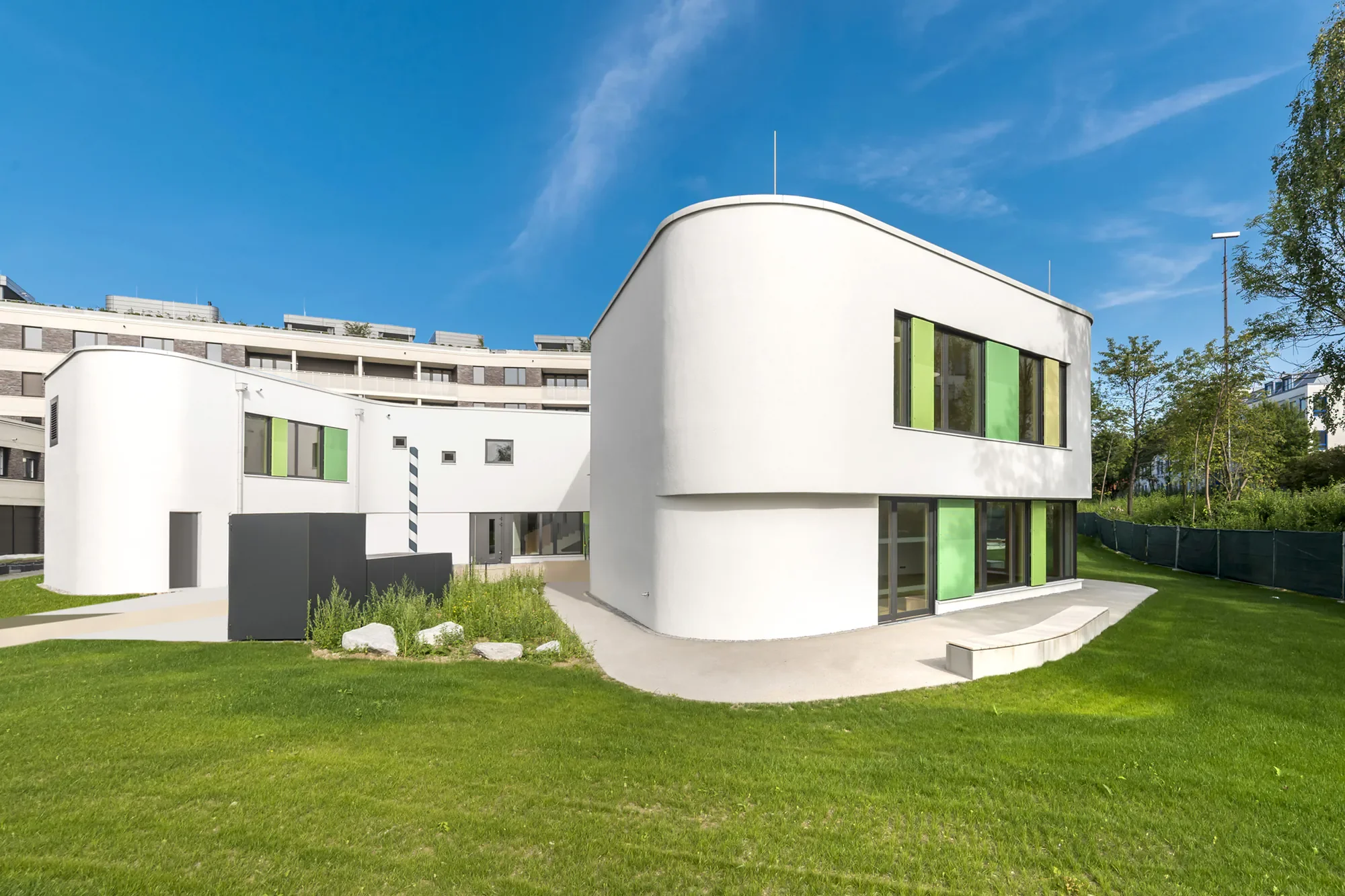 Modernes weißes Wohngebäude mit großen Fenstern und grünen Akzenten, umgeben von grünem Rasen und Bäumen, unter blauem Himmel.