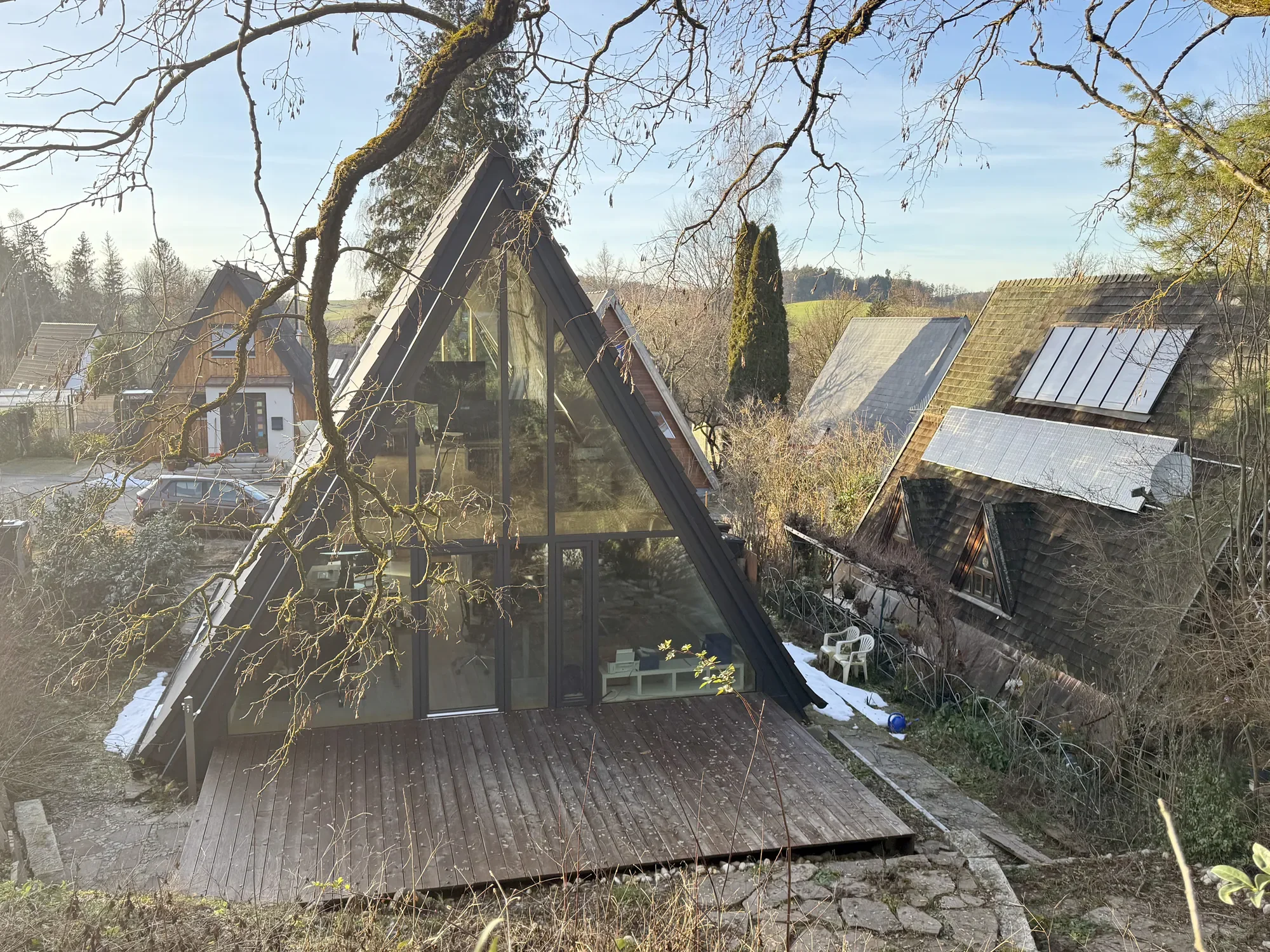Ein Haus mit einem großen, dreieckigen Glasfenster. Es befindet sich ein Holzboden vor der Tür und es gibt einen Garten mit Bäumen und einem Sitzbereich.