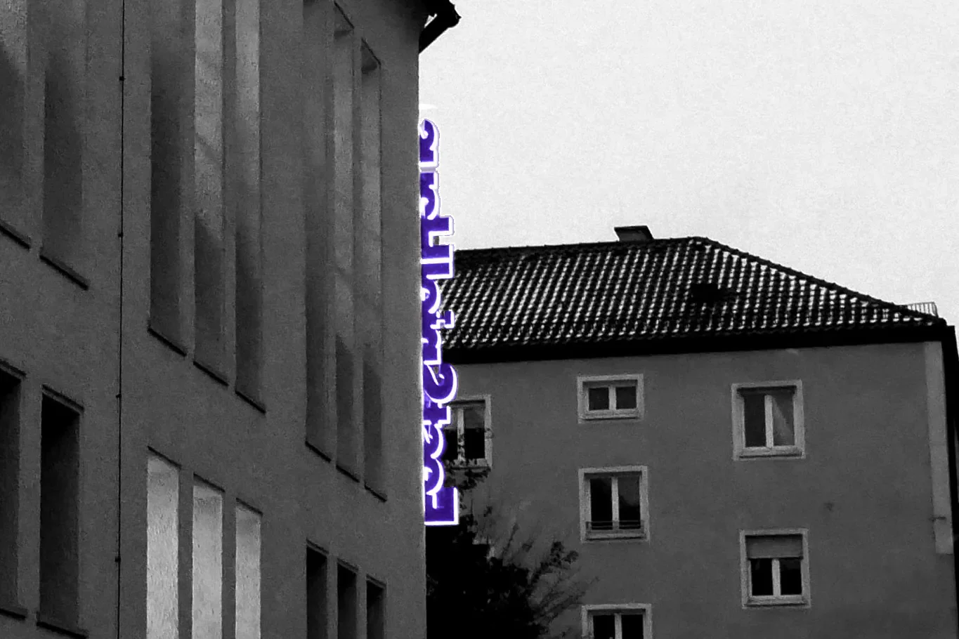 Zwei Mehrfamilienhäuser bei Nacht, eines mit vertikalen Fenstern, das andere mit kleinen rechteckigen Fenstern, und ein vertikaler neonblauer Schreibwarenschild an der Seite des linken Gebäudes.