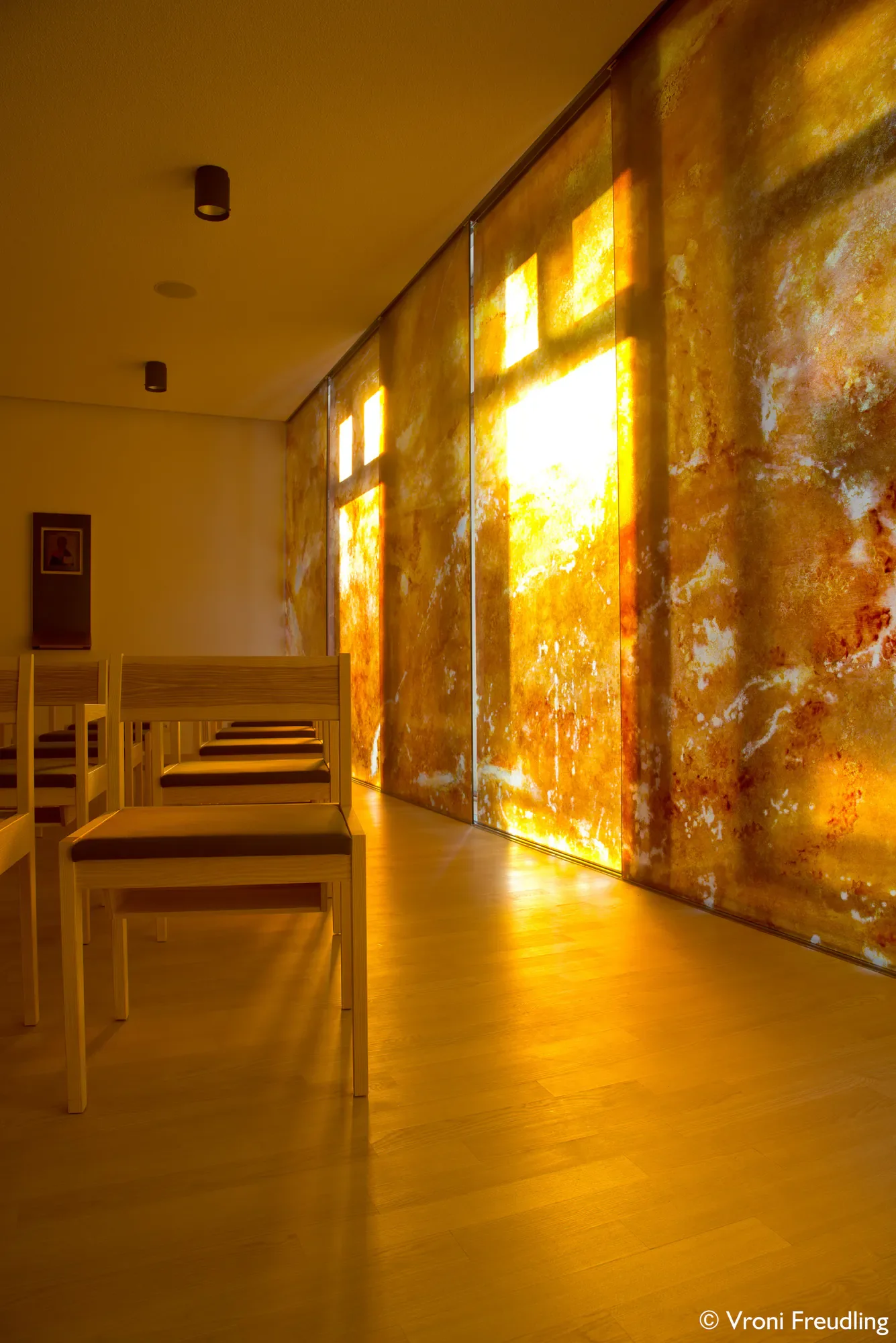 Ein Raum mit Holzstühlen und einer großen hinterleuchteten blattähnlichen Wandinstallation, die an Gold und Orange erinnert, mit Sonnenlicht, das durch Fenster fällt.