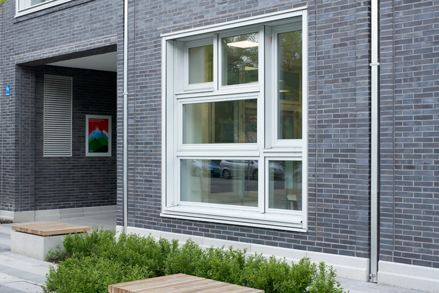 Fenster mit weißen Rahmen an einem modernen Haus mit dunklen Ziegeln, im Vordergrund grüne Pflanzen und Holzdecken