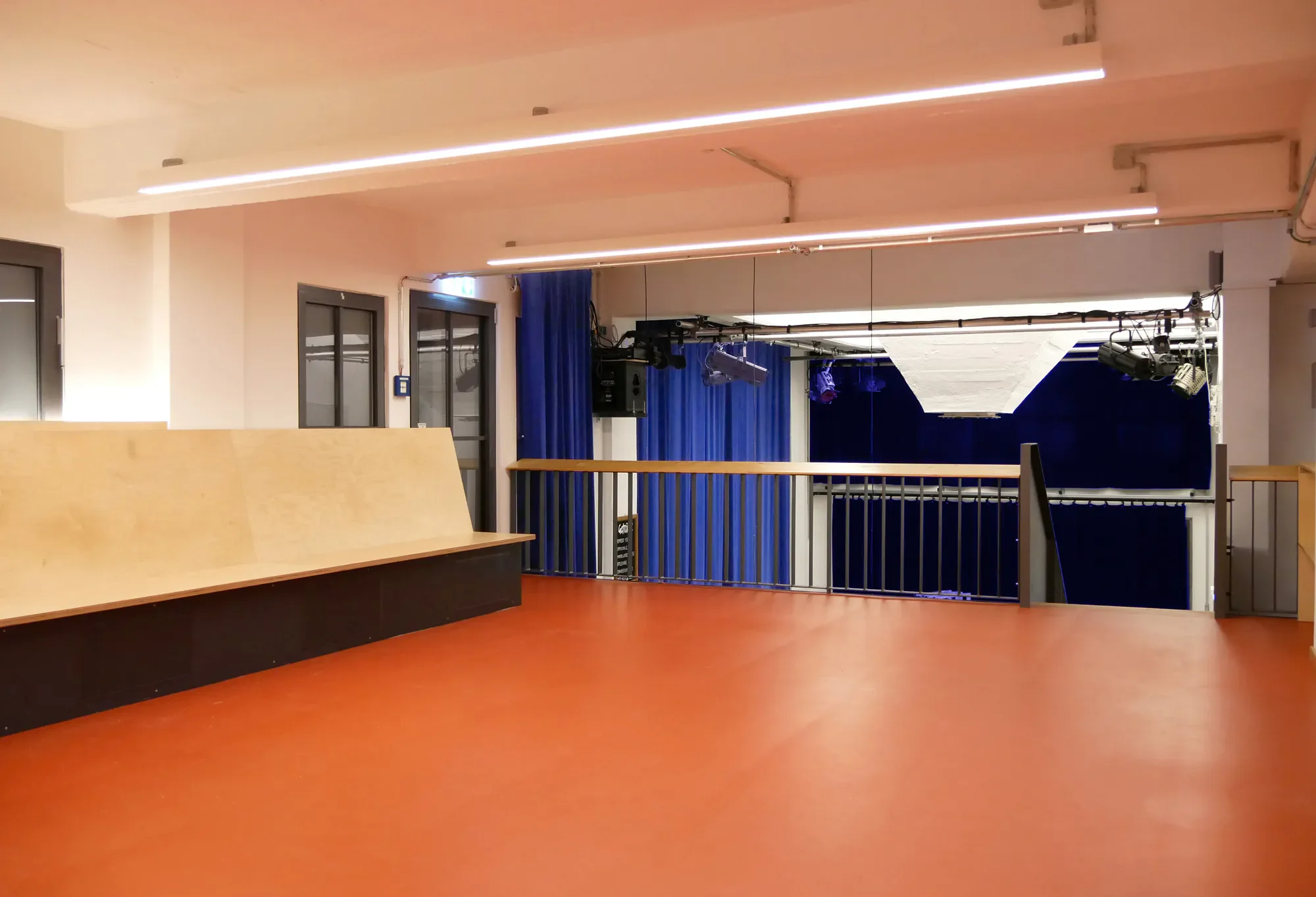 Ein leeres Raum mit roter Boden, weißen Wänden und blauen Vorhängen, eine Barriere und eine kleine Bühne, technische Geräte und Beleuchtung, wahrscheinlich ein Veranstaltungsraum oder Theater.