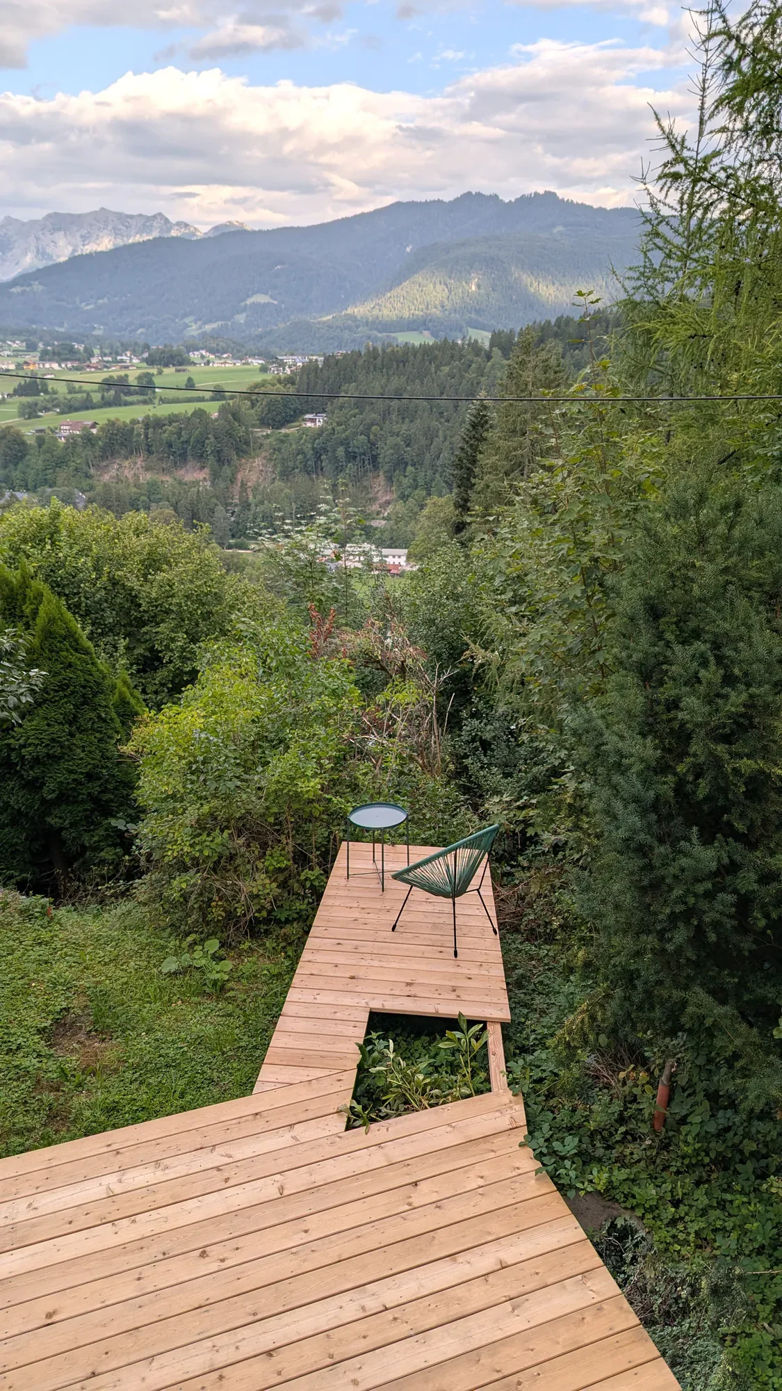Ein Holzdeck mit zwei Stühlen, umgeben von Bäumen und Blick auf eine grüne Berglandschaft mit Bergen im Hintergrund.