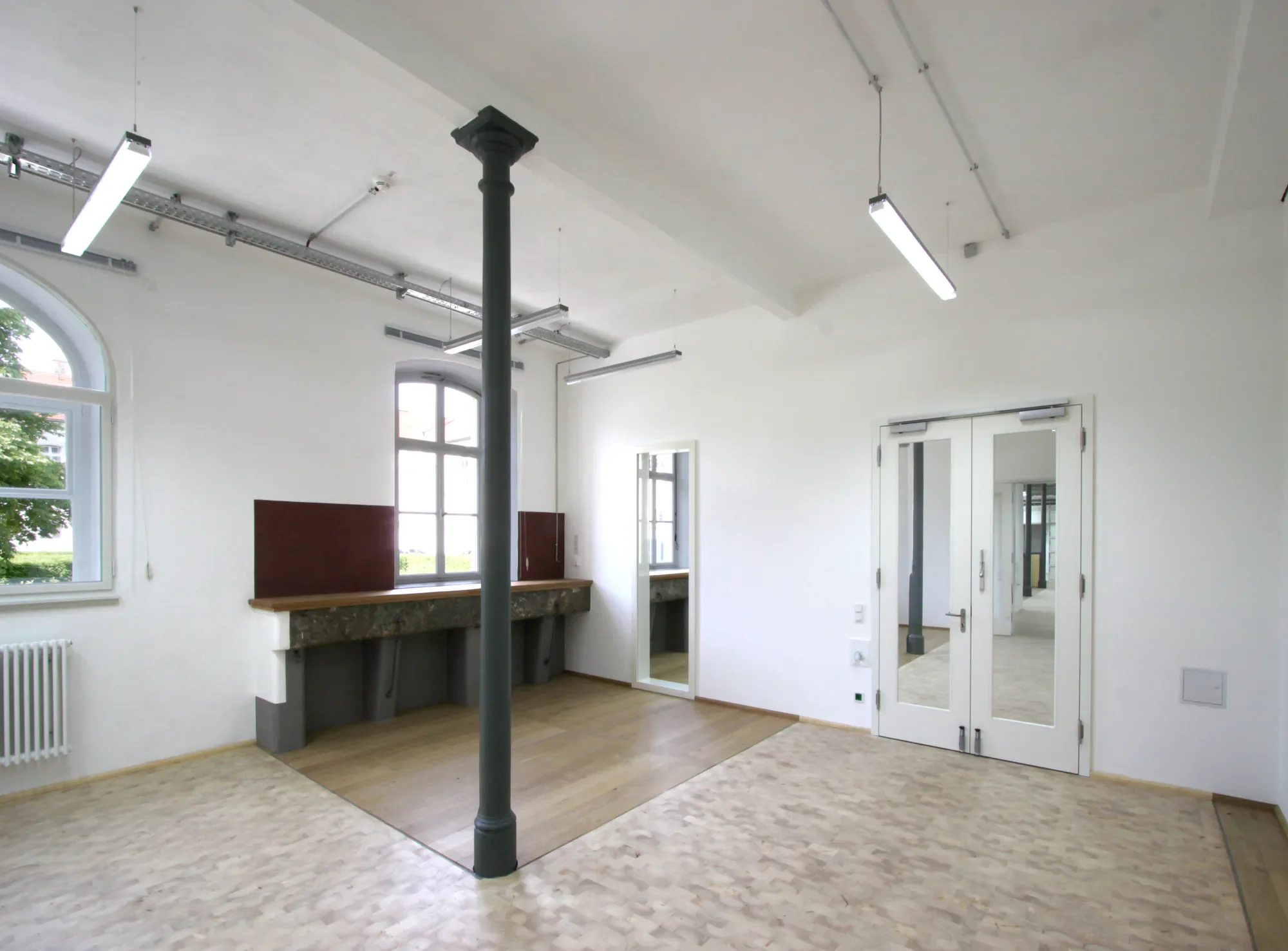 Ein heller Raum mit hohen Decken, großen Fenstern und einer Metallstütze in der Mitte. Es gibt einen kleinen Holzbereich und Türen, die in einen anderen Raum führen.