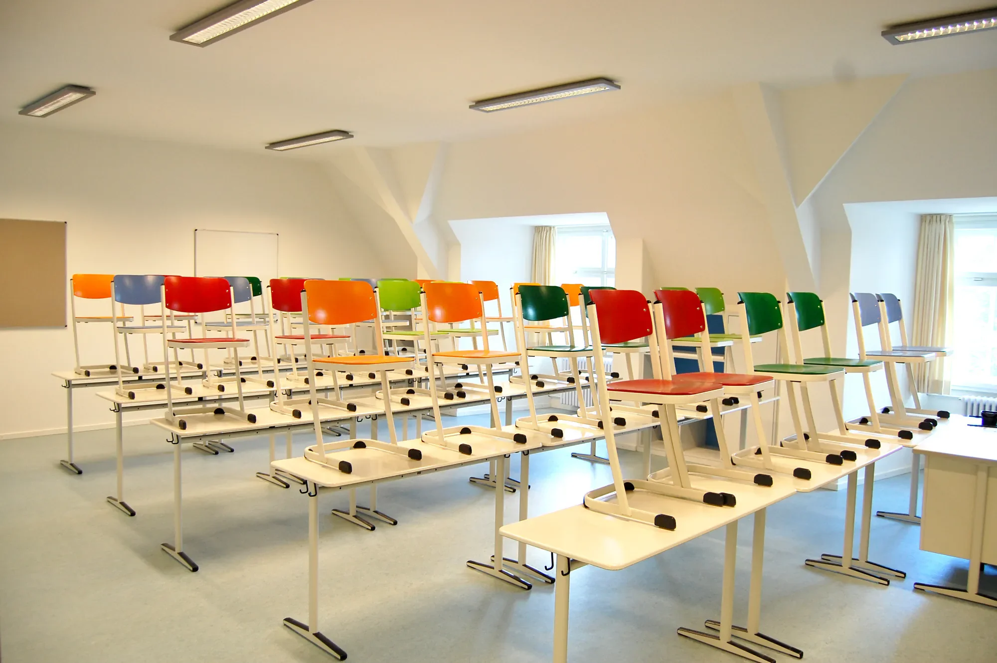 Leere Schulstühle in einem Klassenzimmer, die in mehreren Reihen gestapelt sind, mit bunten Stuhllehnen in Rot, Blau, Grün, Orange und Weiß, vor großen Fenstern mit Vorhängen, darunter eine Tafel.