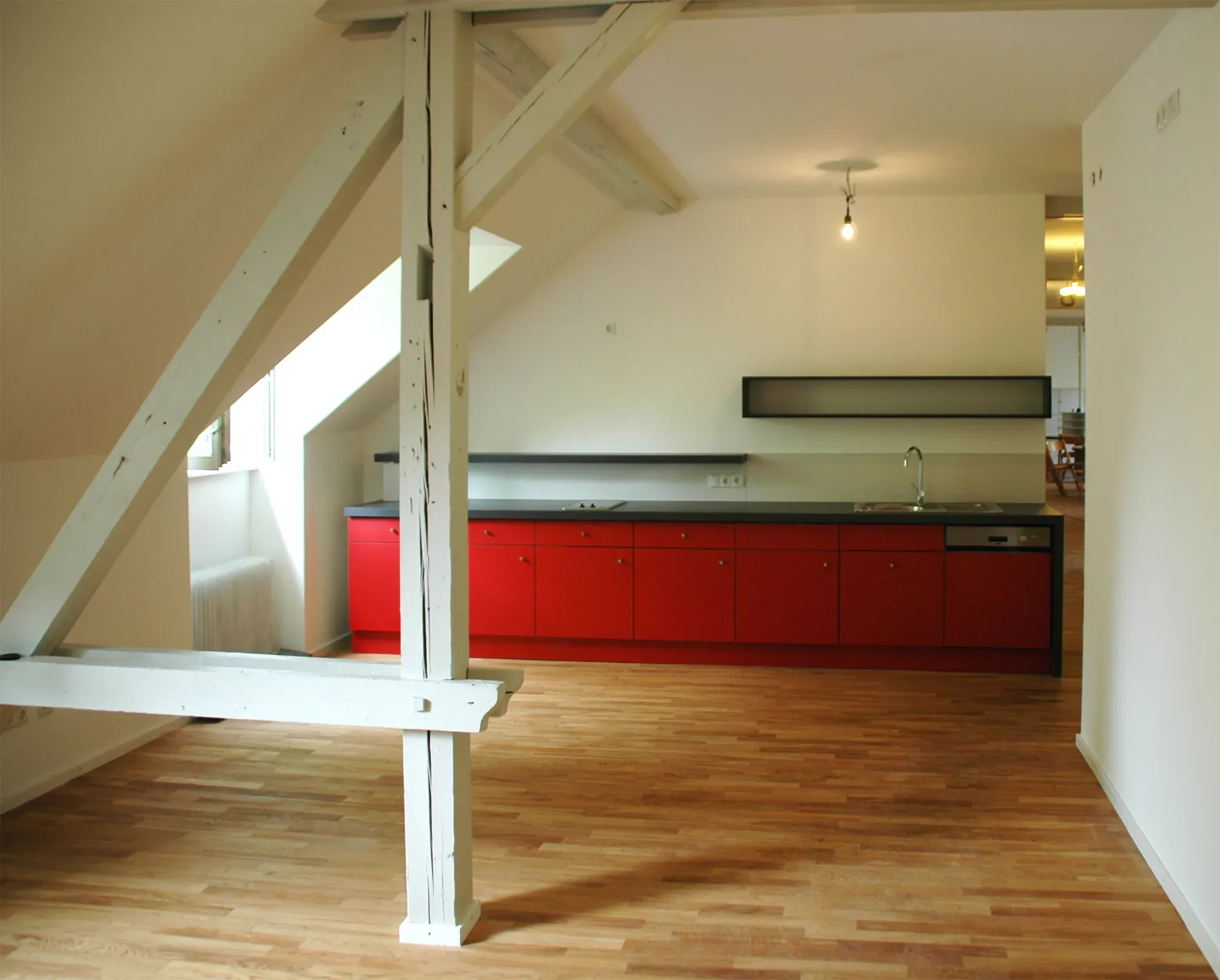 Ein moderner Küchenraum mit roten Schränken, schwarzem Arbeitsplatten, Spüle und Dunstabzug, im Hintergrund eine weiße Wand, große Fenster, Holzboden, offene Raumgestaltung, eine nackte Glühbirne an der Decke, Sicht auf einen Essbereich im Hintergrund.
