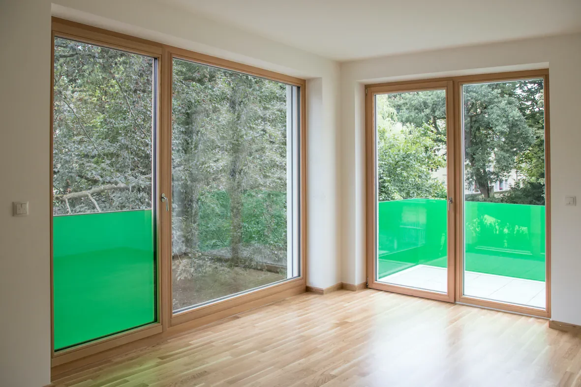Ein leeres Zimmer mit großen Fenstern, durch die man Bäume und ein grünes Balkongeländer sieht.