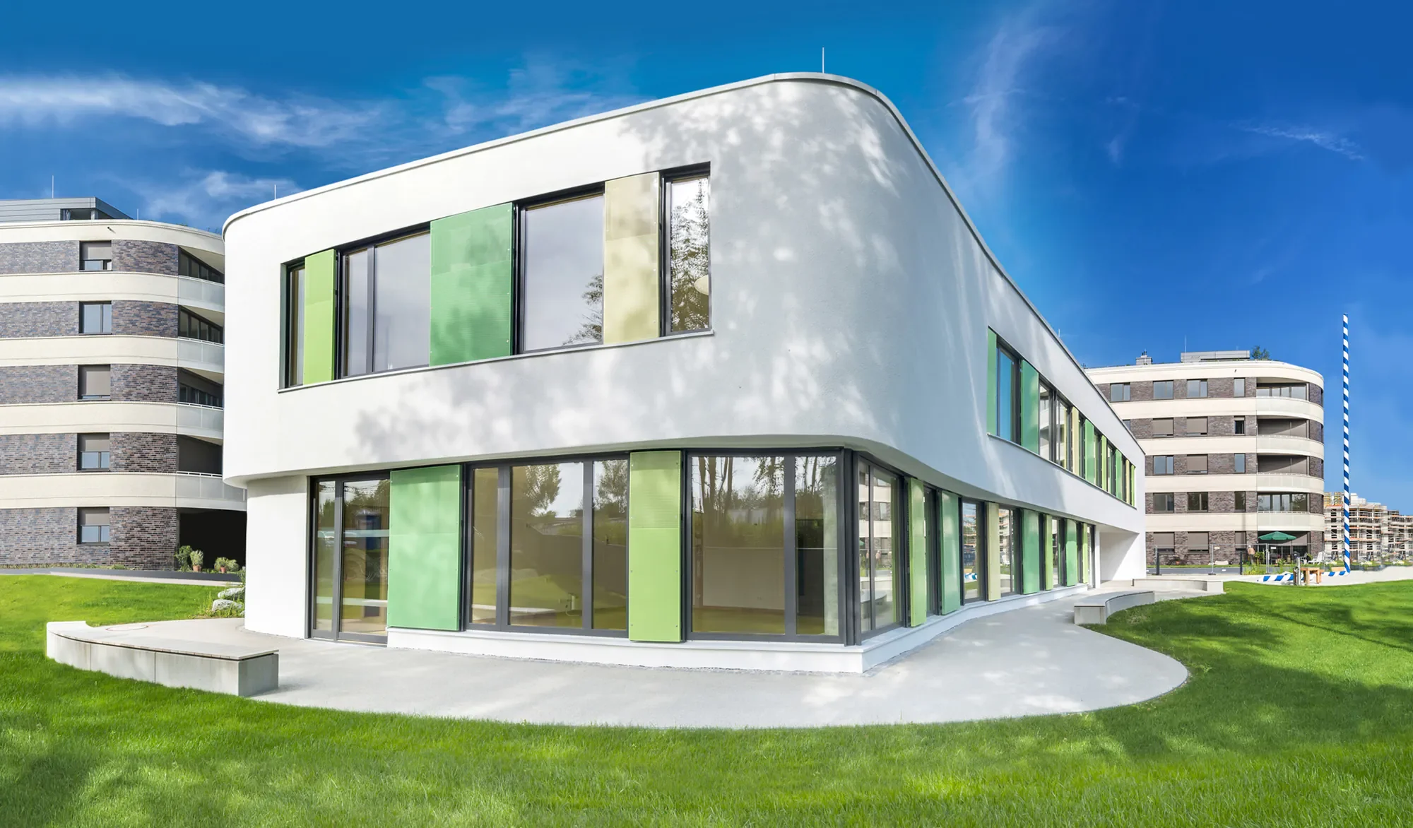 Modernes weißes Gebäude mit großen Fenstern in grünen und gelben Farben, umgeben von grünem Rasen und mehreren weiteren Gebäuden im Hintergrund, bei blauem Himmel mit wenigen Wolken.