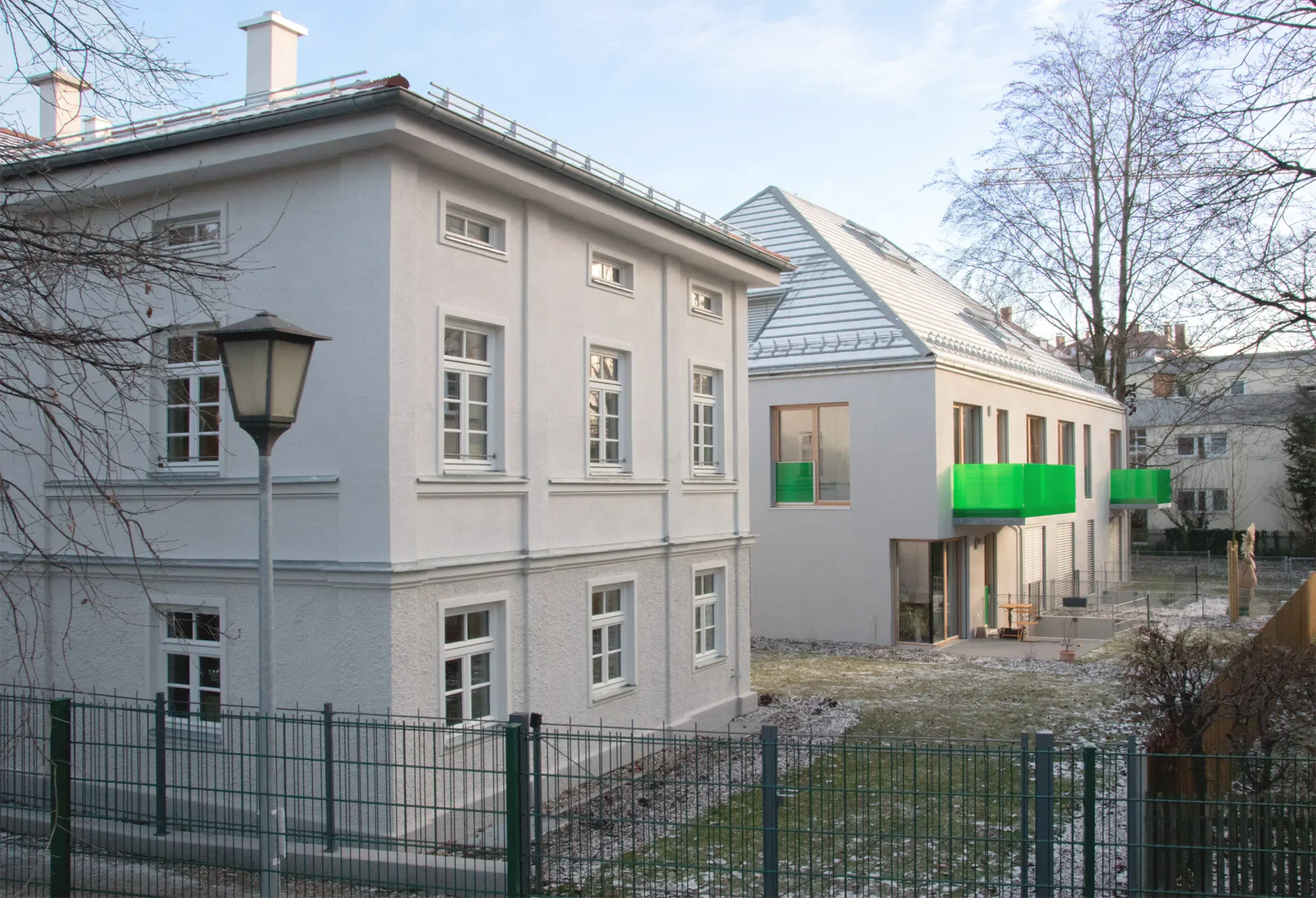 Mehrfamilienhaus mit weißen Wänden, mehreren Fenstern, Balkon mit grüner Verkleidung und einem Garten im Vordergrund, umgeben von einem grünen Zaun, im Hintergrund Bäume und ein blauer Himmel.