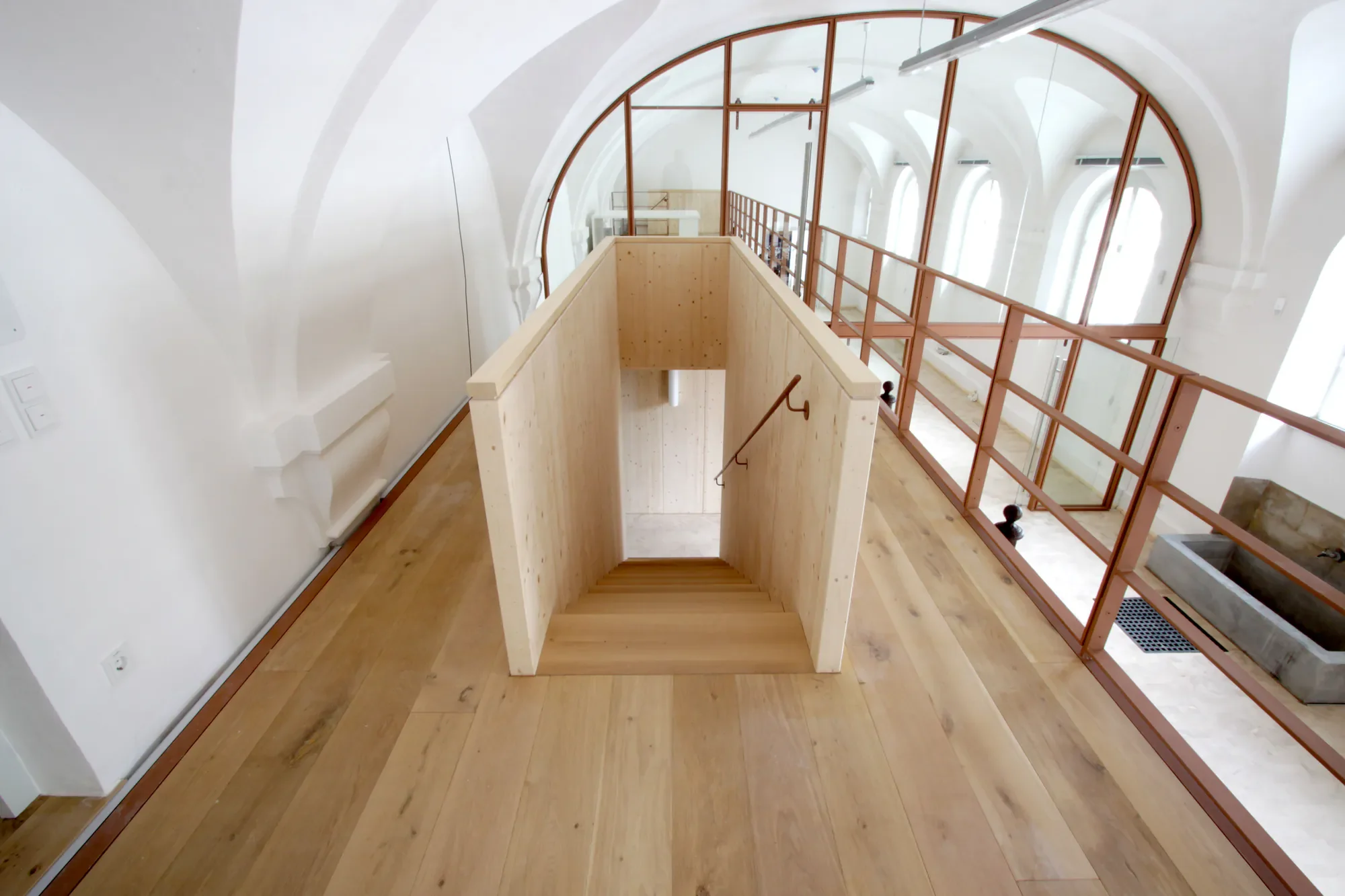 Innenraum eines modernen Gebäudes mit Holztreppe, großen Fenstern und weißer Decke mit gebogenen Linien.