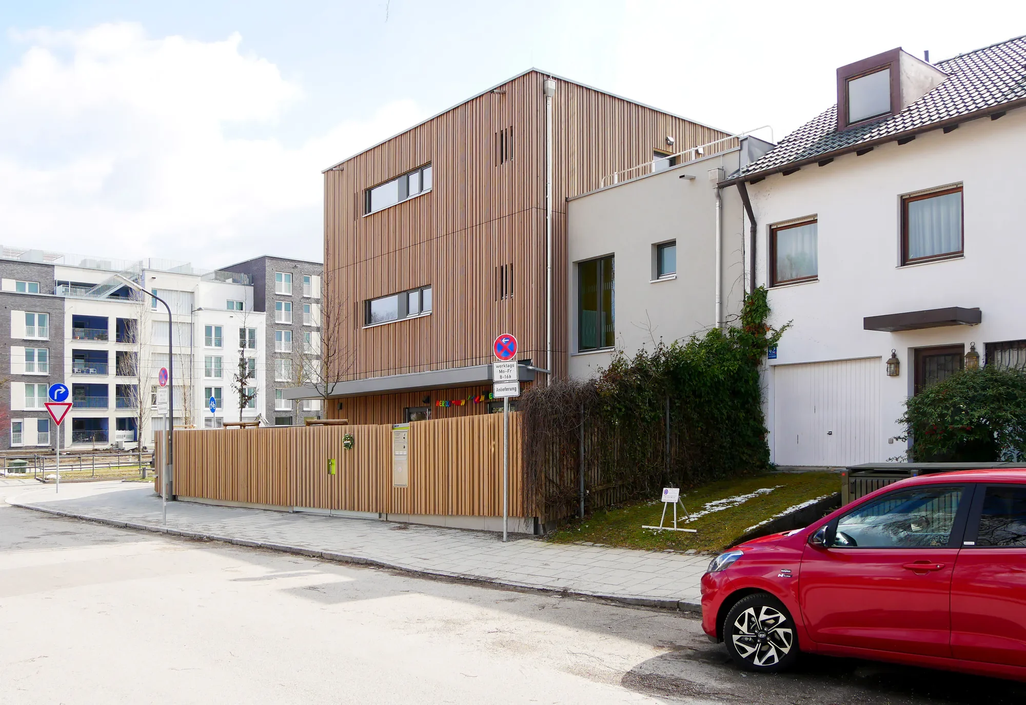 Moderner Gebäudekomplex mit Holz- und Putzfassade, Parkplätze vor dem Haus, E-Scooter Ladestation, Straßenverkehrsschilder, Baum und Gartenbereich im Vordergrund.