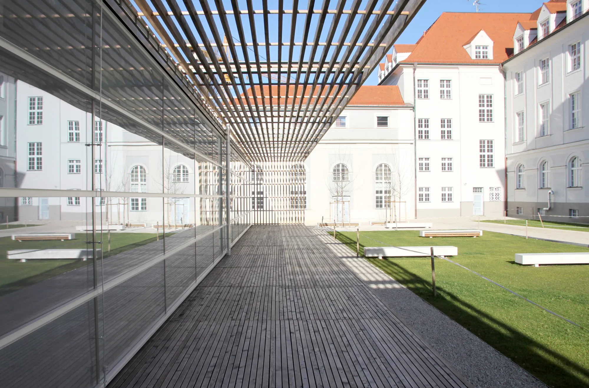 Moderner Gebäudekomplex mit Holz- und Glasarchitektur vor einem historischen weißen Gebäude mit roten Ziegeldächern, sonniger Tag, kahle Bäume im Hof.