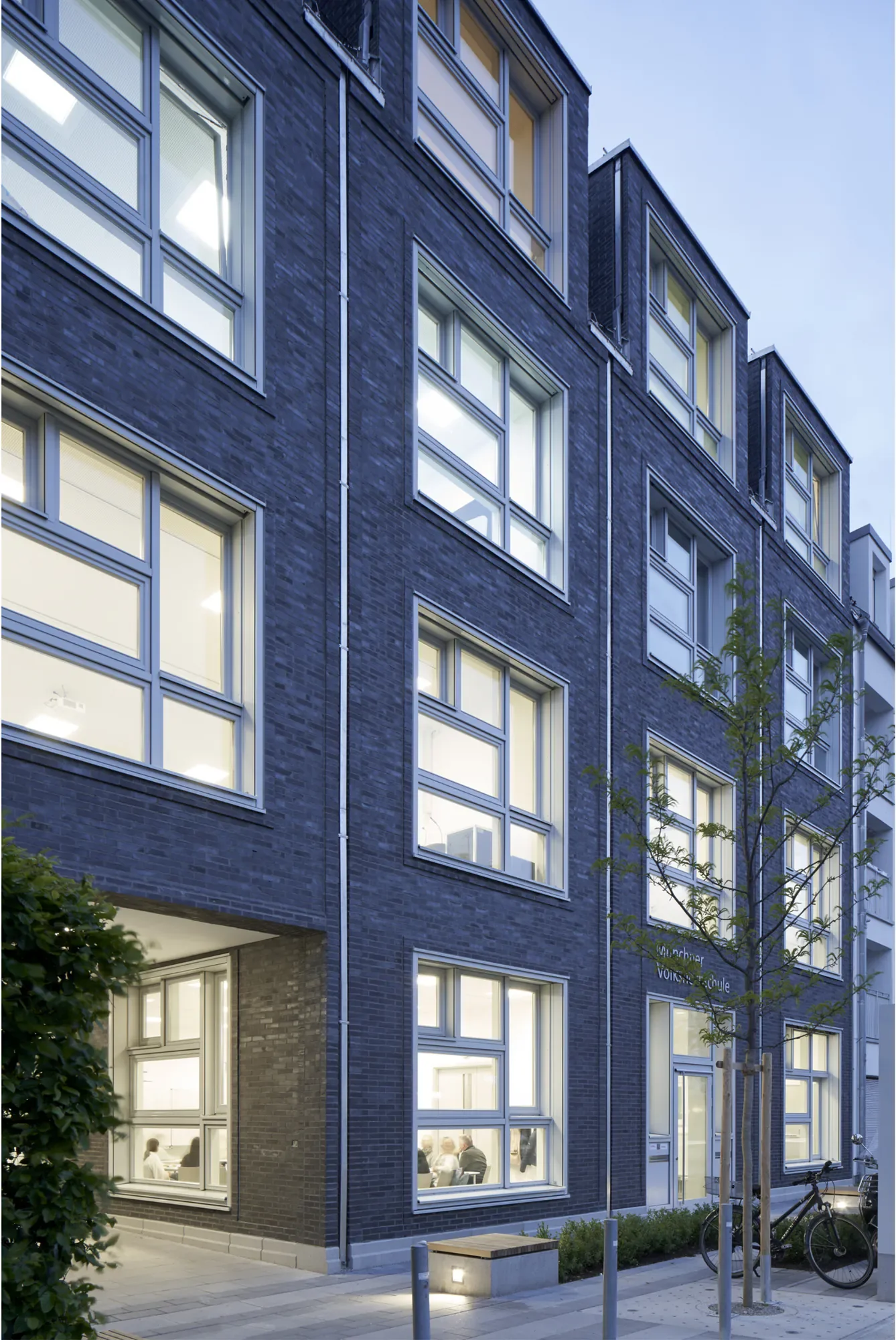 Modernes mehrstöckiges Gebäude mit großen Fenster, einige sind beleuchtet, und einem gepflegten Gehweg mit Fahrradständern, Bäumen und Straßenlaternen