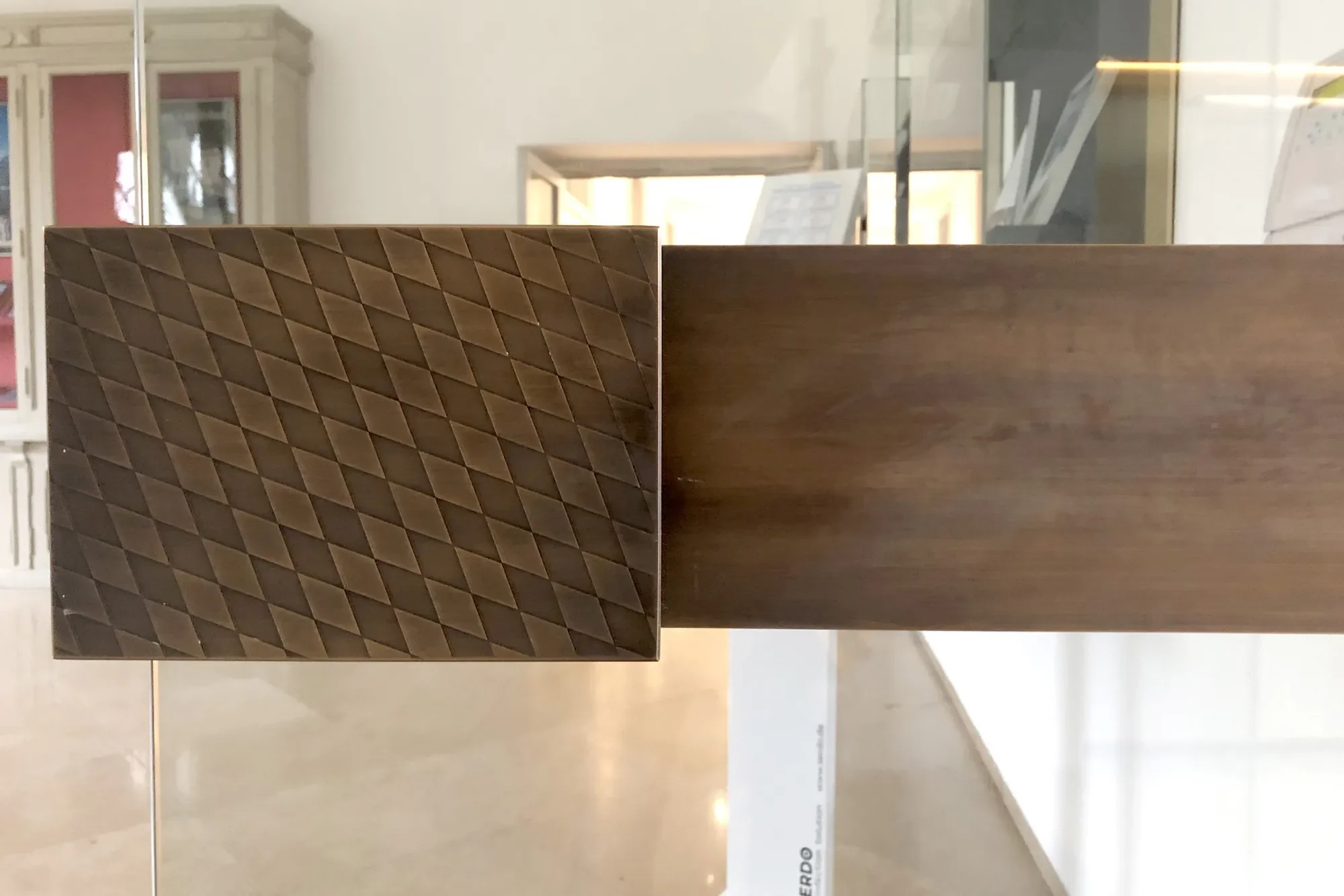 Ein Ausstellungsstück mit einer dekorativen, geprägten Holzoberfläche auf der linken Seite, und eine glatte, etwas dunklere Holzfläche auf der rechten Seite, im Inneren eines Museums oder Ladens, mit Glas und anderen Objekten im Hintergrund.