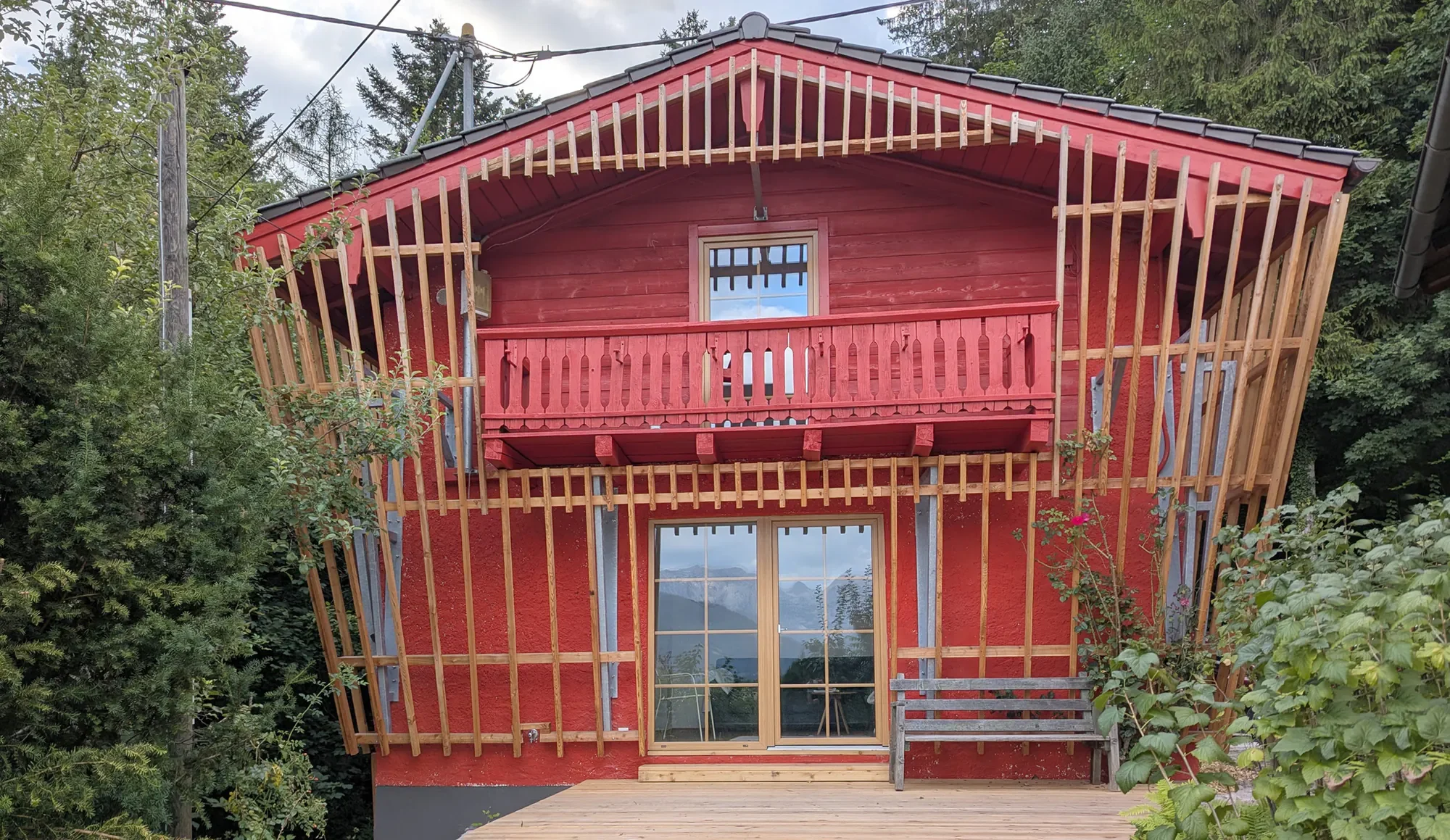 Haus im Bau mit roter Wand, Holzrahmen für Dacharbeiten und Balkon, umgeben von Bäumen.