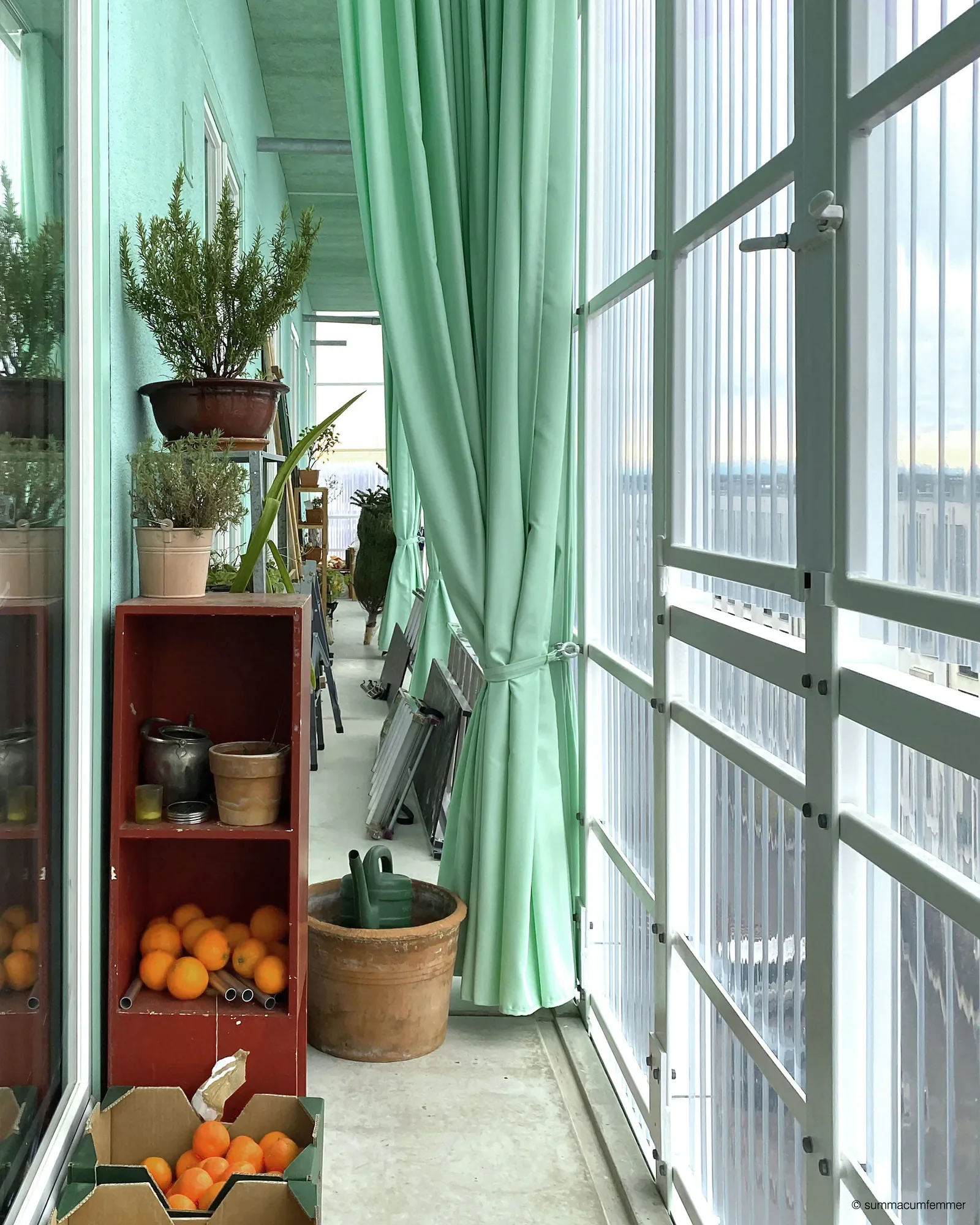 Blick auf einen Balkon mit grünen Vorhängen, Blumenkübeln und Kisten voller Orangen, sowie einem Wasserhahn an der Wand