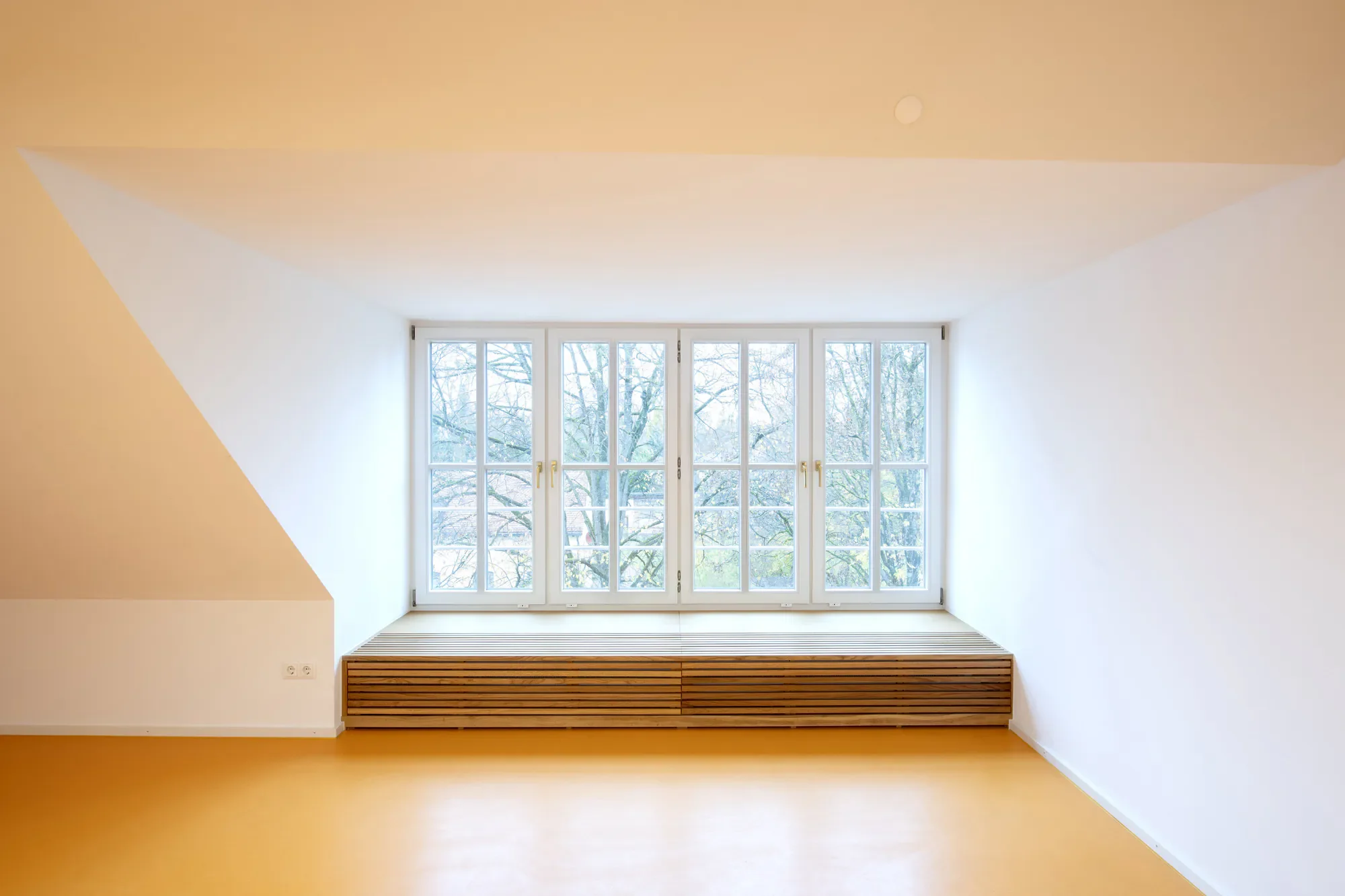 Ein heller, moderner Raum mit einer großen Fensterfront, die nach draußen auf Bäume blickt, und einem Holzbank-Element vor den Fenstern.