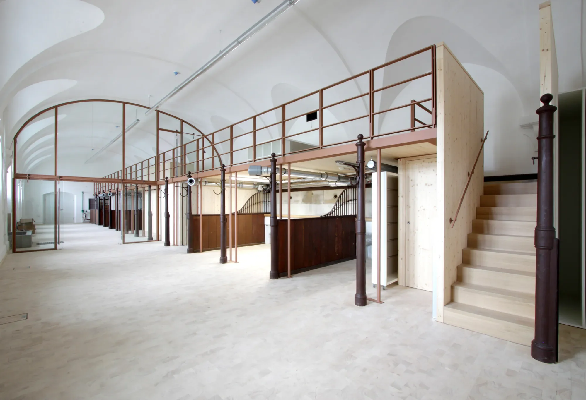 Innenansicht eines leeren, modernen Gebäudes mit hohem, gewölbtem Decken, einer Treppe aus Holz und umlaufendem Stahlgeländer sowie mehreren braunen Eisenpfählen.