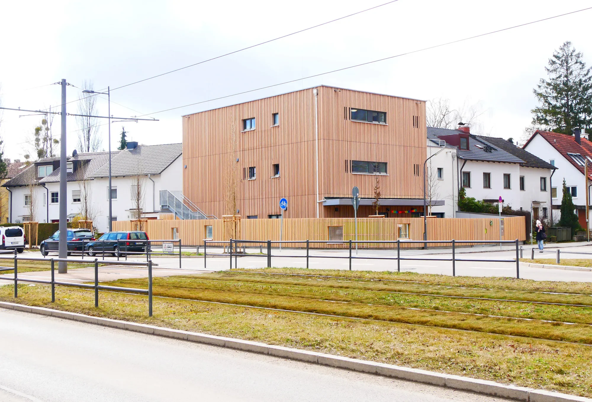 Ein modernes, mehrstöckiges Holzgebäude inmitten eines Wohngebietes, umgeben von traditionellen Einfamilienhäusern, mit einer Fahrradspur im Vordergrund und parkenden Autos.