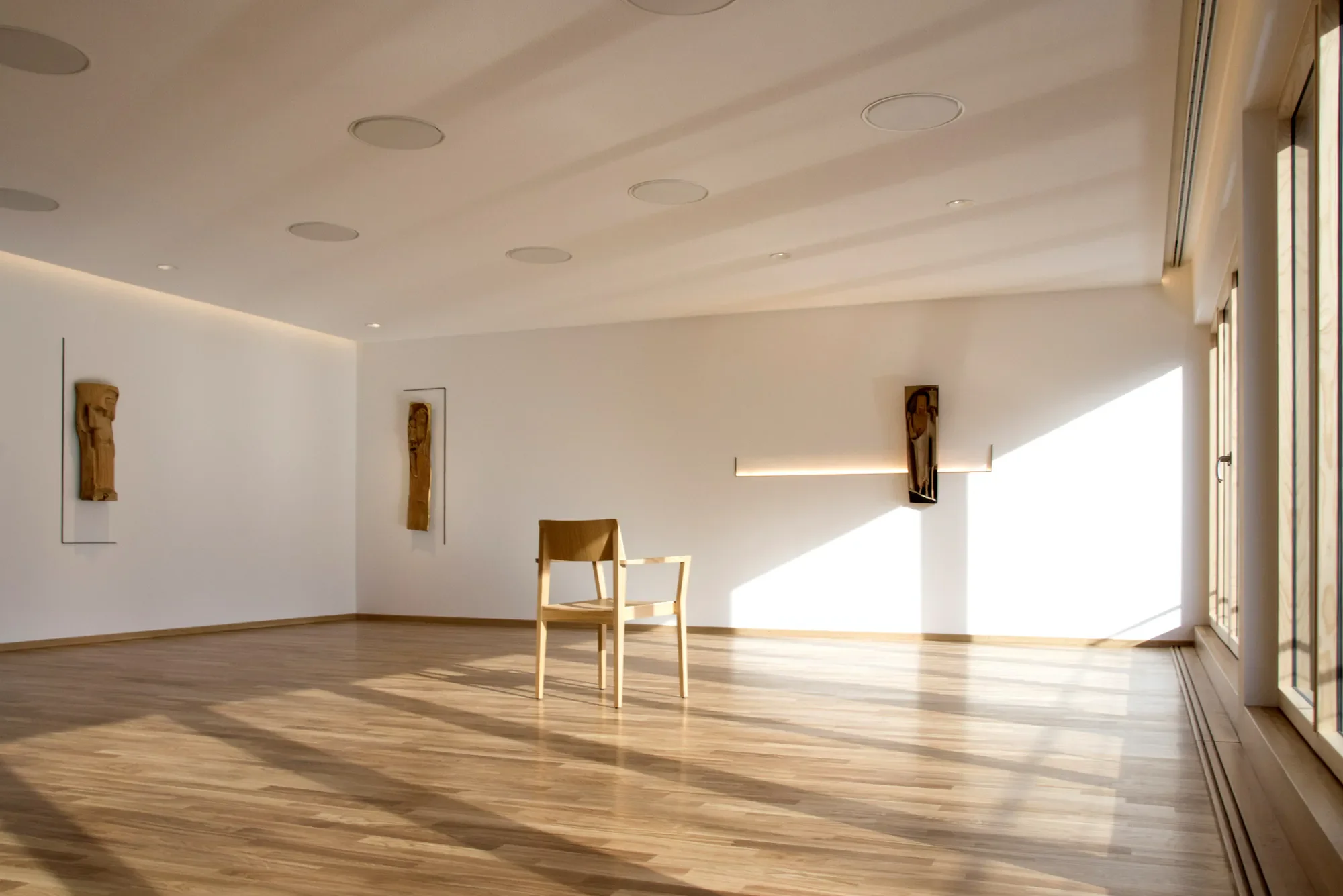 Ein helles, modernes Wohnzimmer mit Holzboden, weißen Wänden und mehreren kreisförmigen Deckenleuchten. Es gibt Kunstwerke an den Wänden und eine große Fensteröffnung auf der rechten Seite, die Sonnenlicht hineinlässt.
