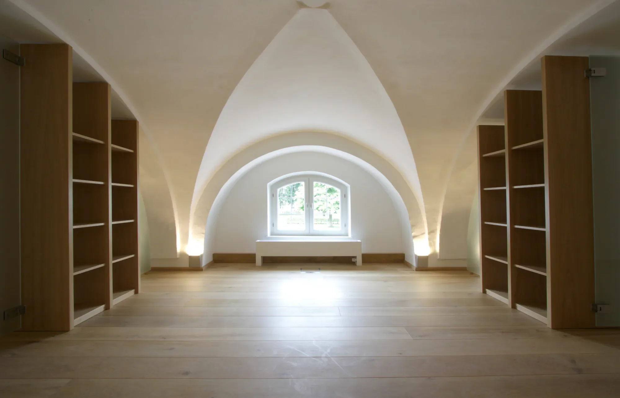 Innenansicht eines Zimmers mit hohen, gewölbten Decken und einer kleinen Fensteröffnung, eingerahmt von weißen Wänden und Holzboden, flankiert von leeren Bücherregalen auf beiden Seiten.