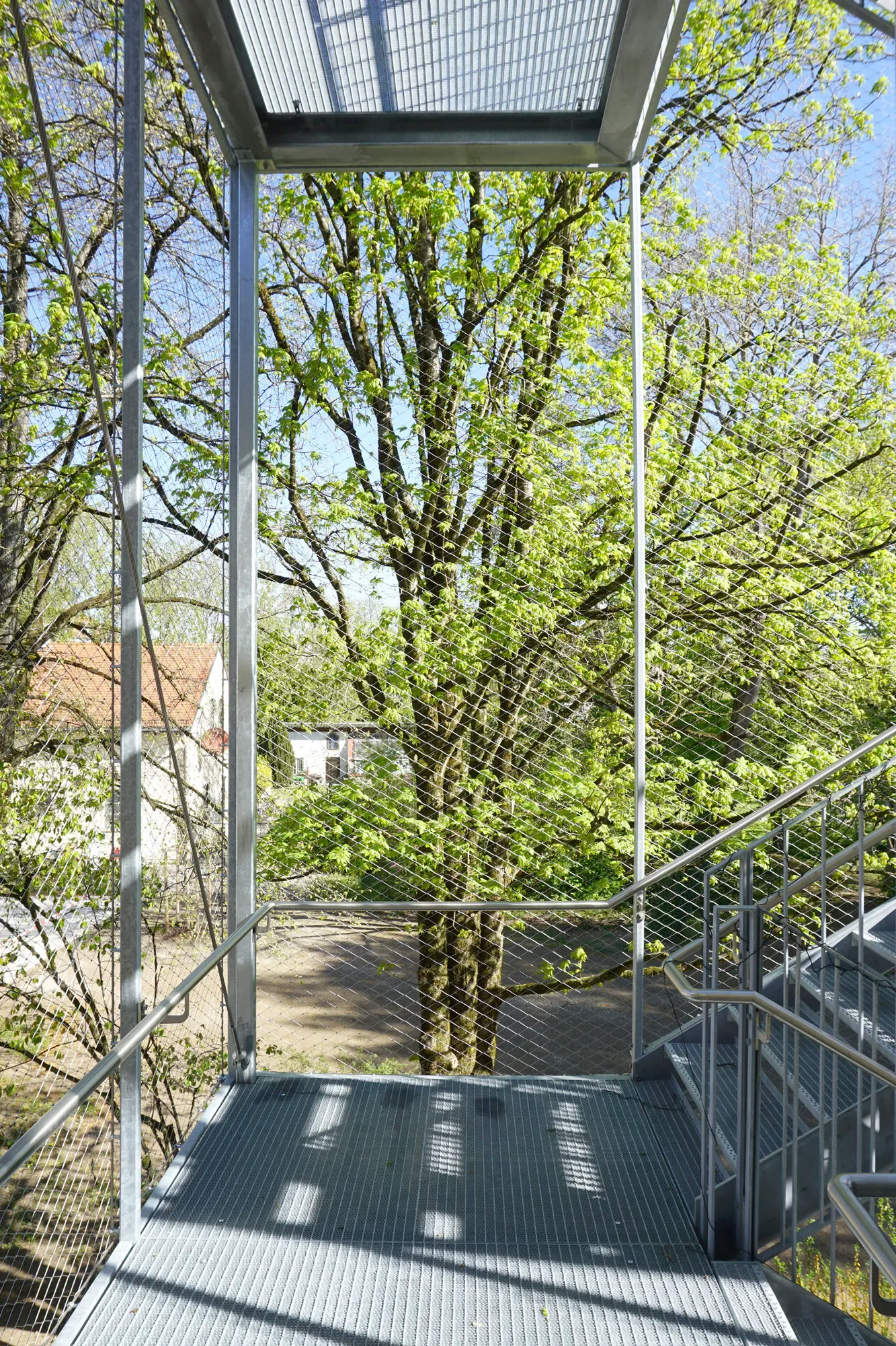 Blick von einer Metalltreppe mit Geländer auf einen grünen Baum mit lichtgrünen Blättern und blauen Himmel im Hintergrund.