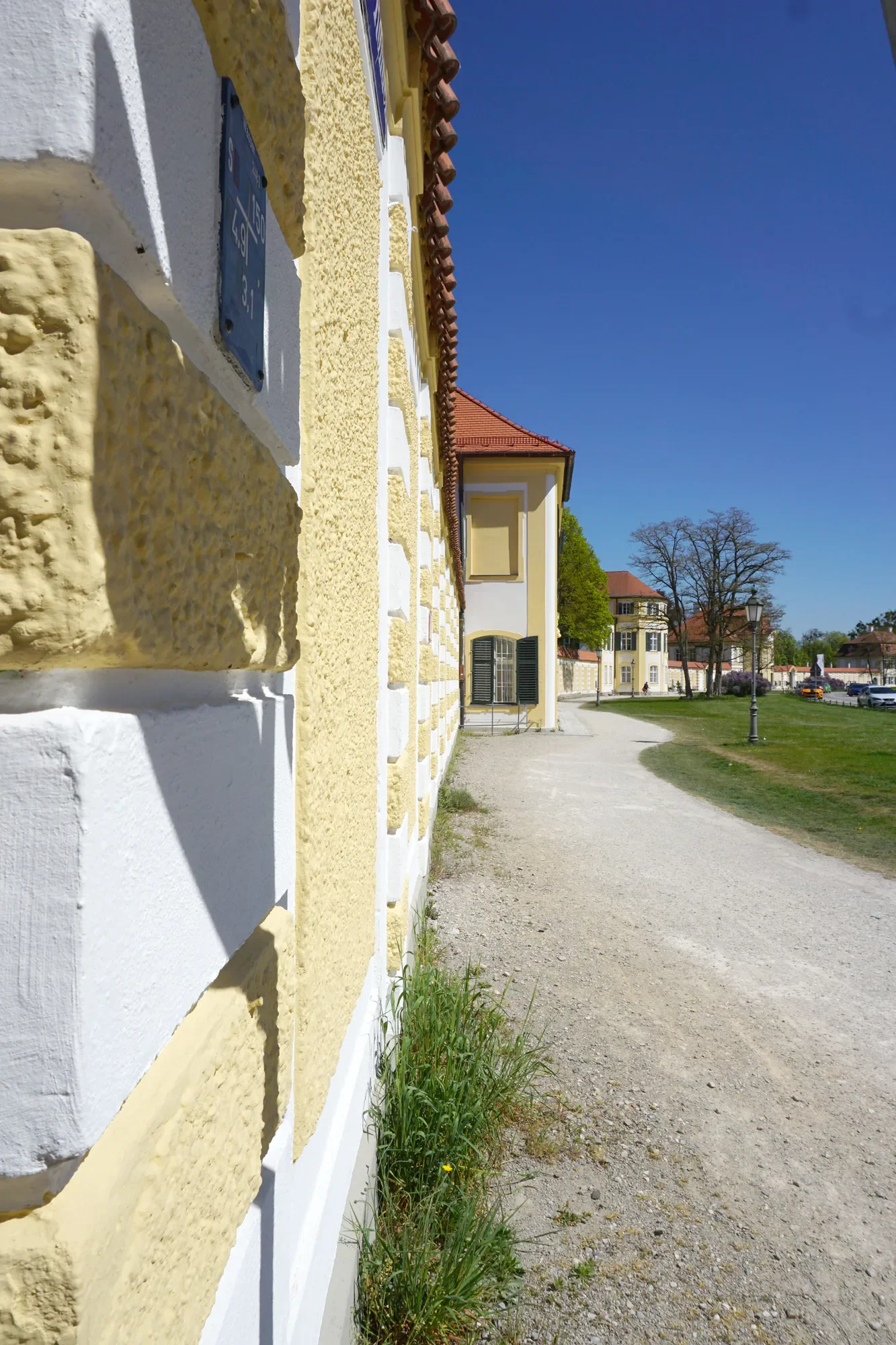 Ein gelb-weißes Gebäude mit roten Dachziegeln. Es gibt Fenster mit geschlossenen schwarzen Jalousien und einen Weg entlang. Im Hintergrund sind Bäume und andere Gebäude unter klarem blauen Himmel.