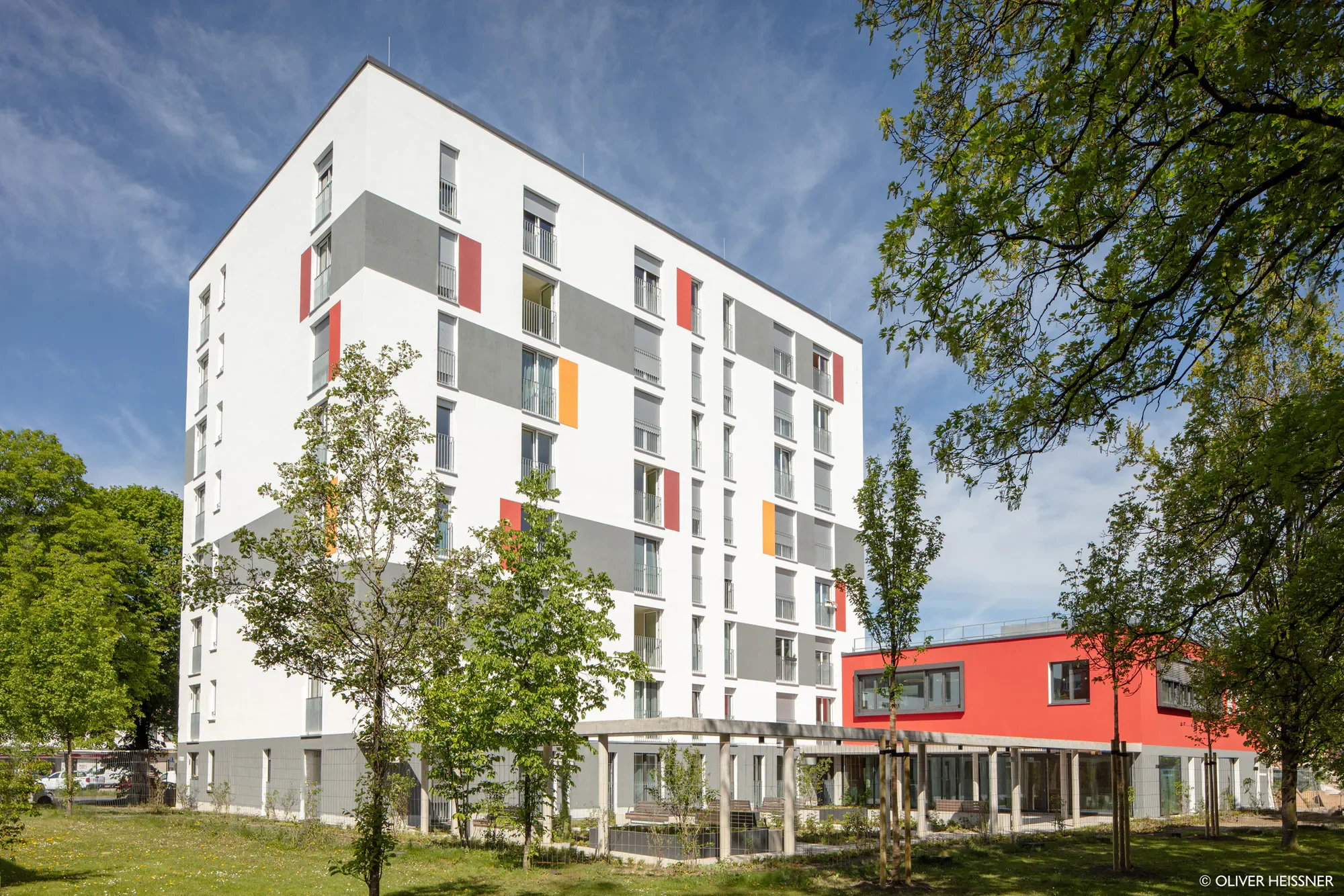 Ein modernes mehrstöckiges Wohngebäude mit weißen, grauen und roten Fassaden, umgeben von Bäumen auf einem grünen Rasen, bei schönem Himmel.