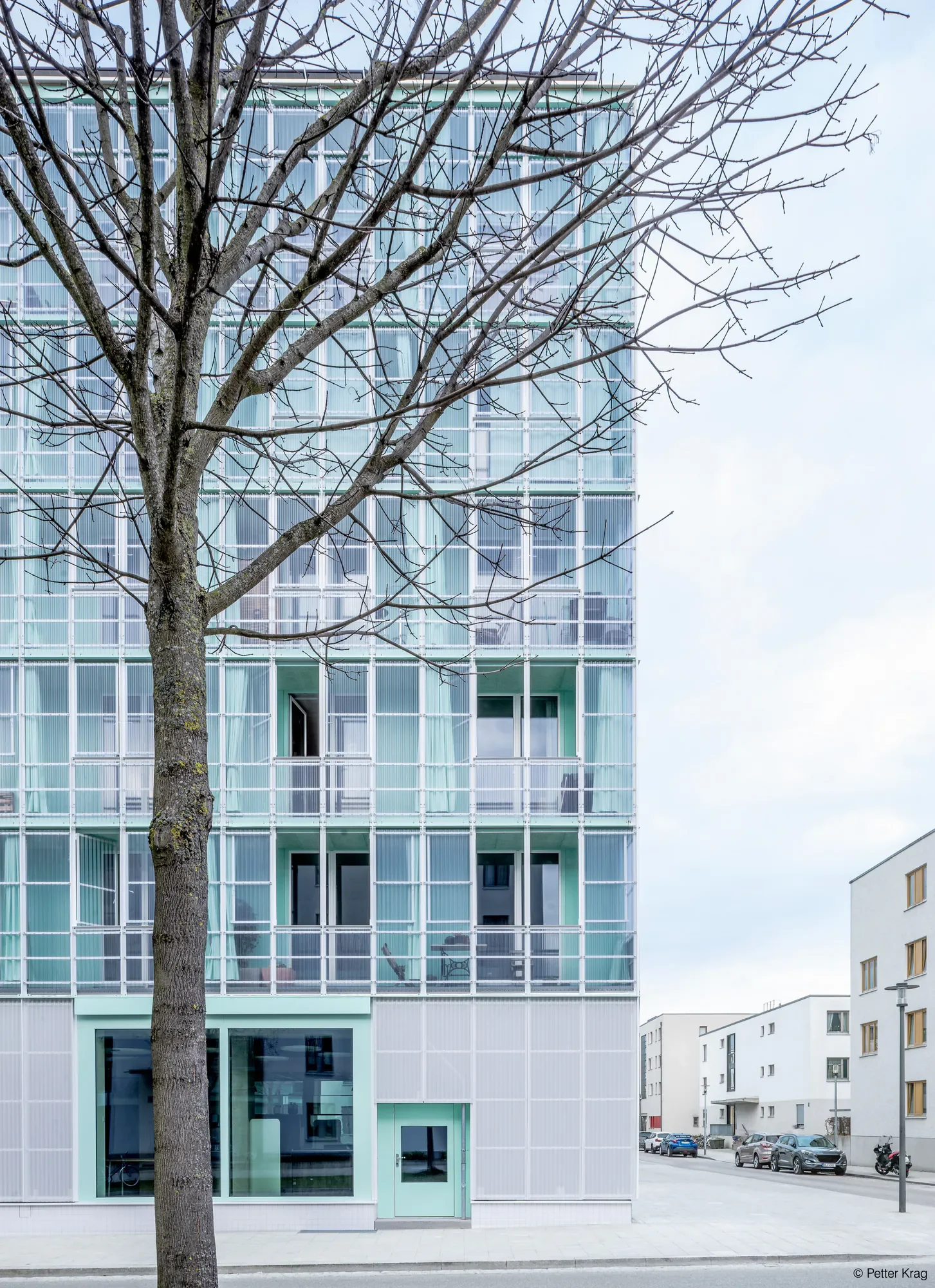Ein moderner Gebäudekomplex mit großen Fenstern, im Vordergrund ein Baum ohne Blätter und im Hintergrund mehrere parkende Autos auf einem Parkplatz.