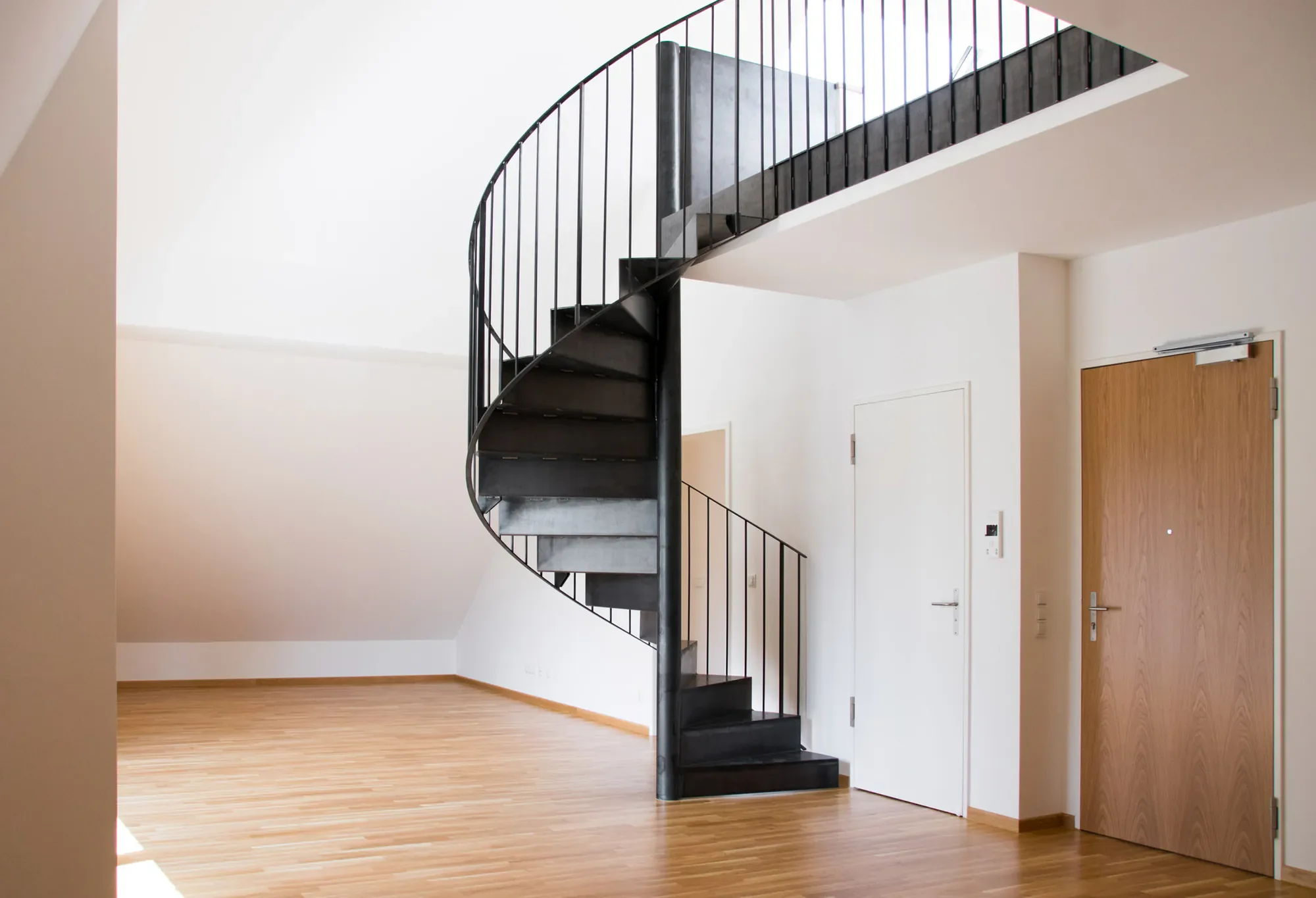 Innenansicht eines modernen Hauses mit schwarzer Spiraltreppe, Holzfußboden und weißen Wänden. Es gibt eine Tür mit Holzoptik auf der rechten Seite.