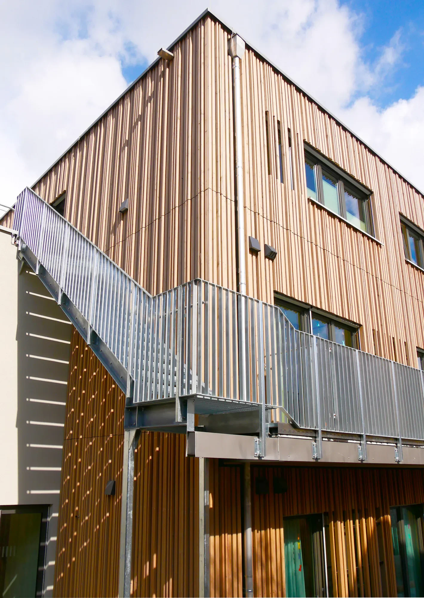 Modernes Gebäude mit Holzfassade und Metall-Außentreppe, blaue Himmel mit Wolken im Hintergrund.