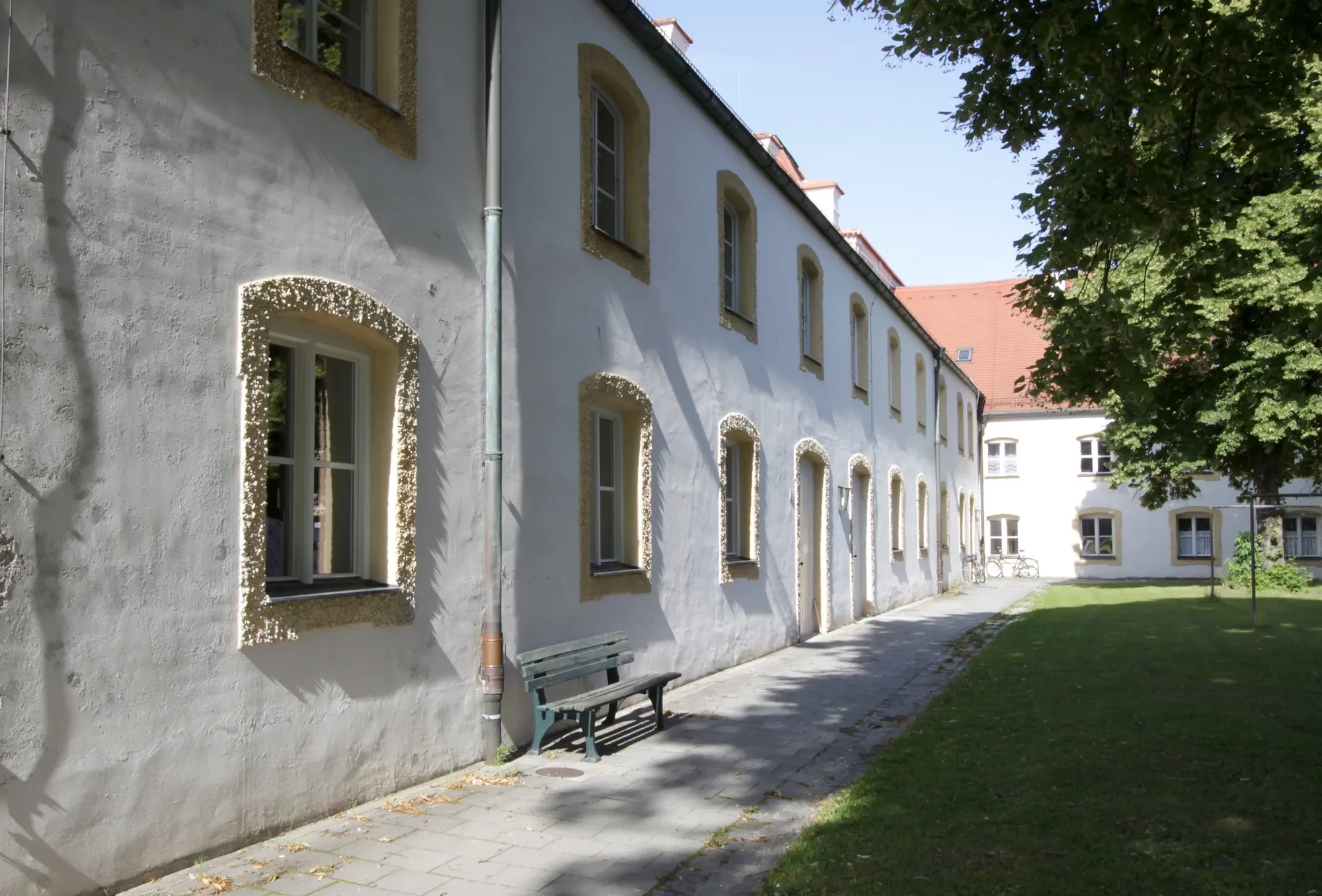 Ein weißes Mehrfamilienhaus mit mehreren kleinen, rechteckigen Fenstern und einem roten Ziegeldach. Es ist von Bäumen umgeben, und es gibt einen Schatten im Vordergrund, der auf den Gehweg fällt.