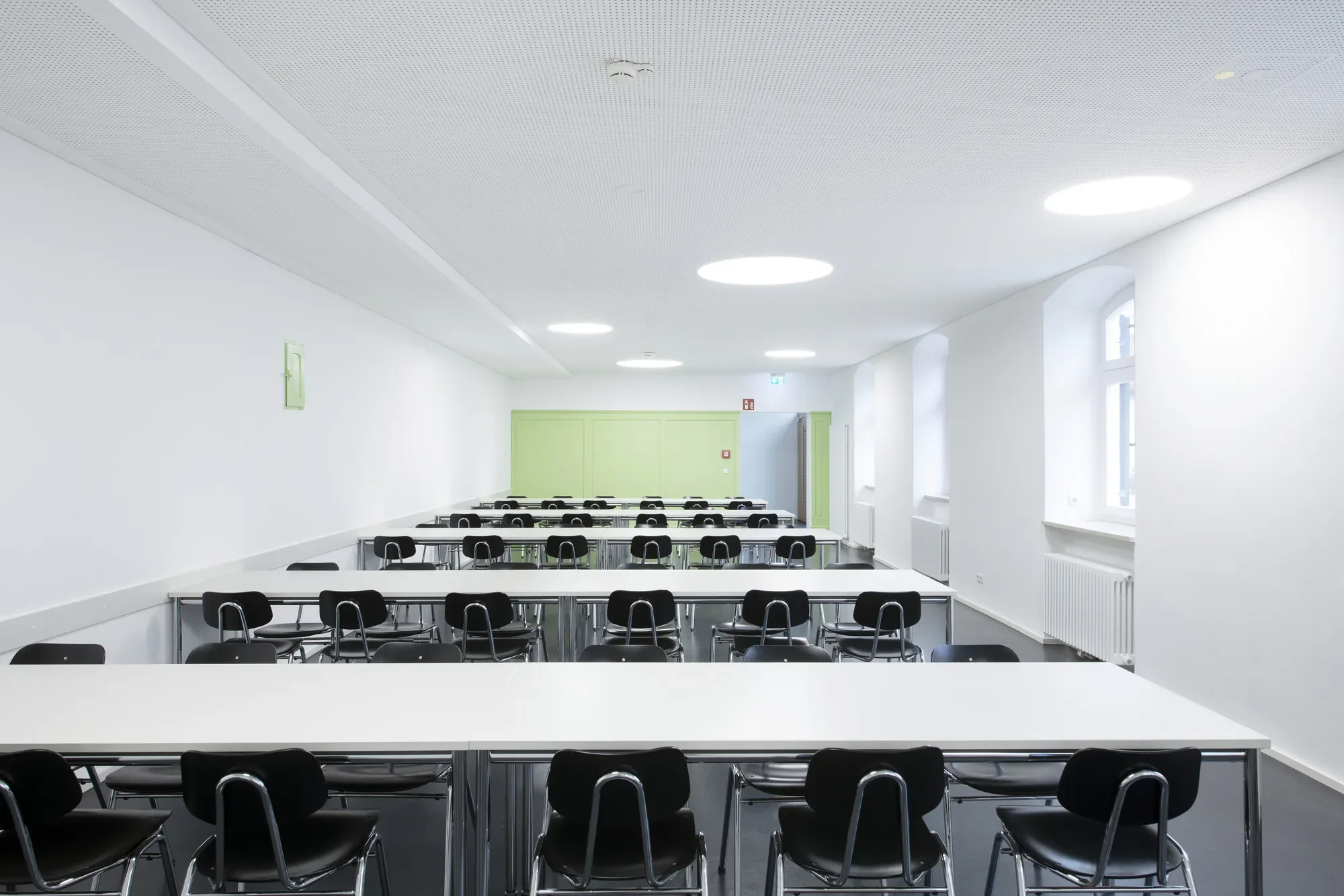 Ein heller, moderner Klassenraum mit weißen Wänden, mehreren Reihen von schwarzen Stühlen und Tischen, großen Fenstern und einer grünen Tür im Hintergrund, Deckenbeleuchtung und Heizkörper an den Wänden.