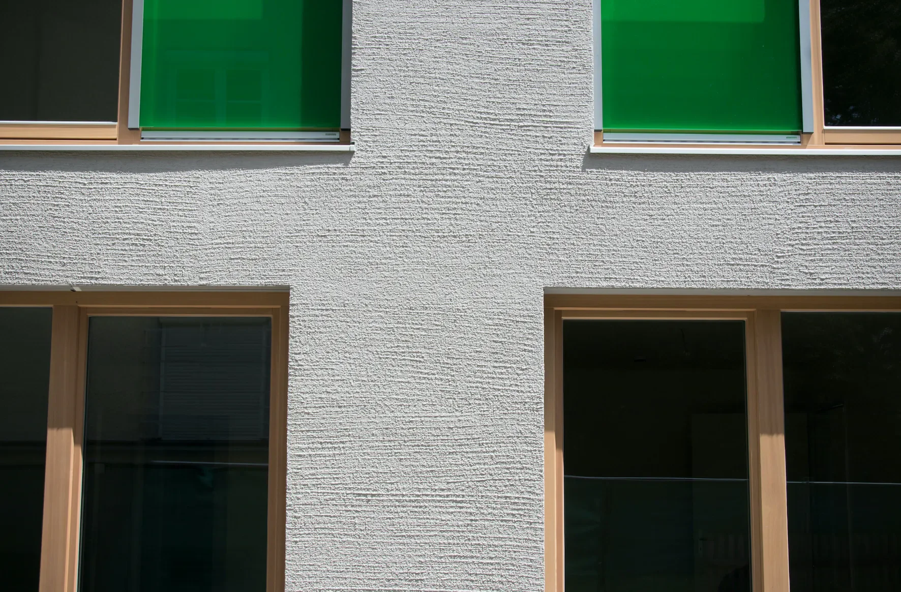 Ein modernes Gebäude mit weißer, strukturierter Außenfassade und großen Fenstern mit Holzrahmen, zwei obere Fenster mit grünen Blenden.