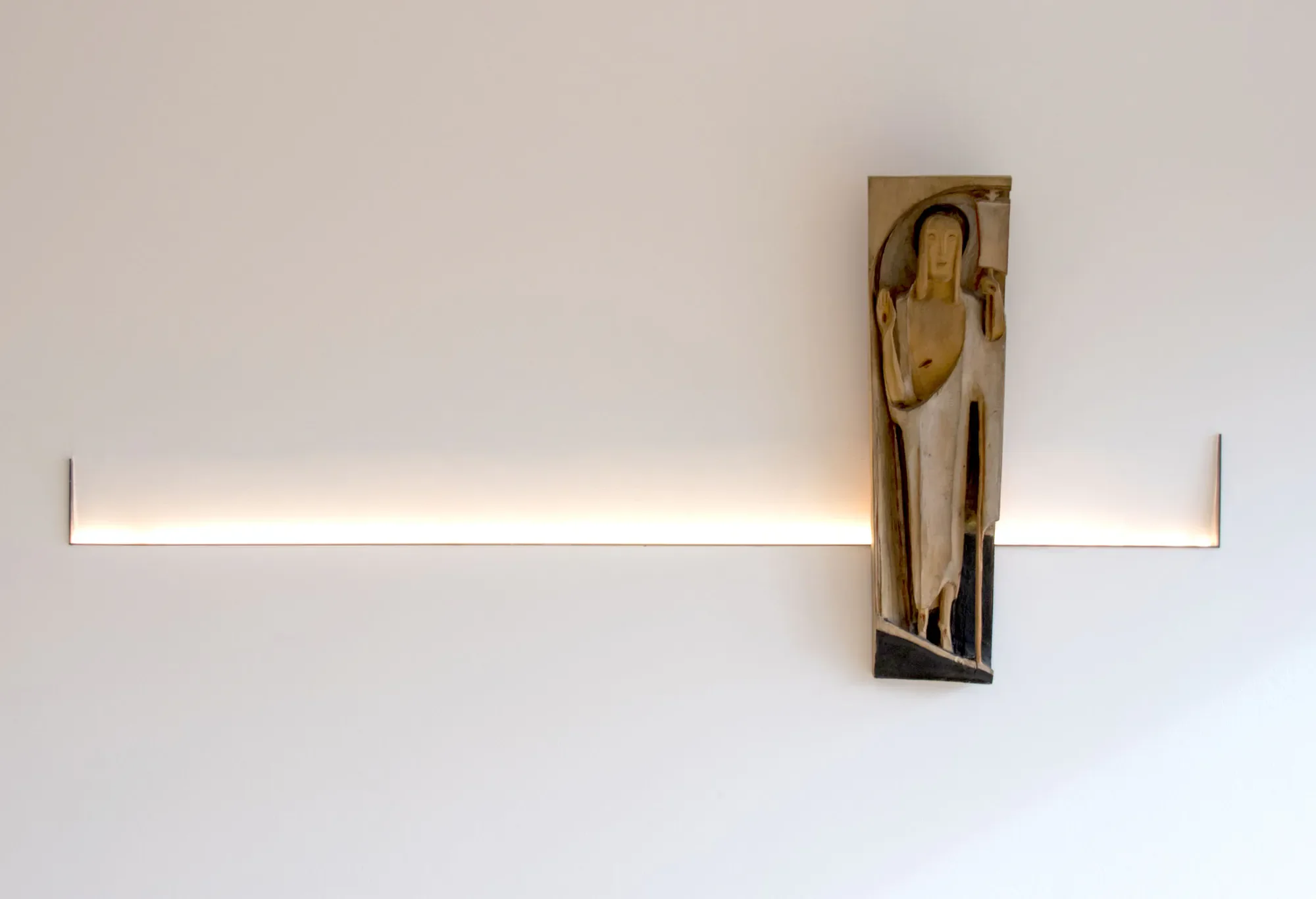 Ein moderner beige Badezimmerraum mit indirekter Beleuchtung an der unteren Wandkante, eine bemalte Holzskulptur an der Wand, die einen menschlichen Kopf und Schulter zeigt.