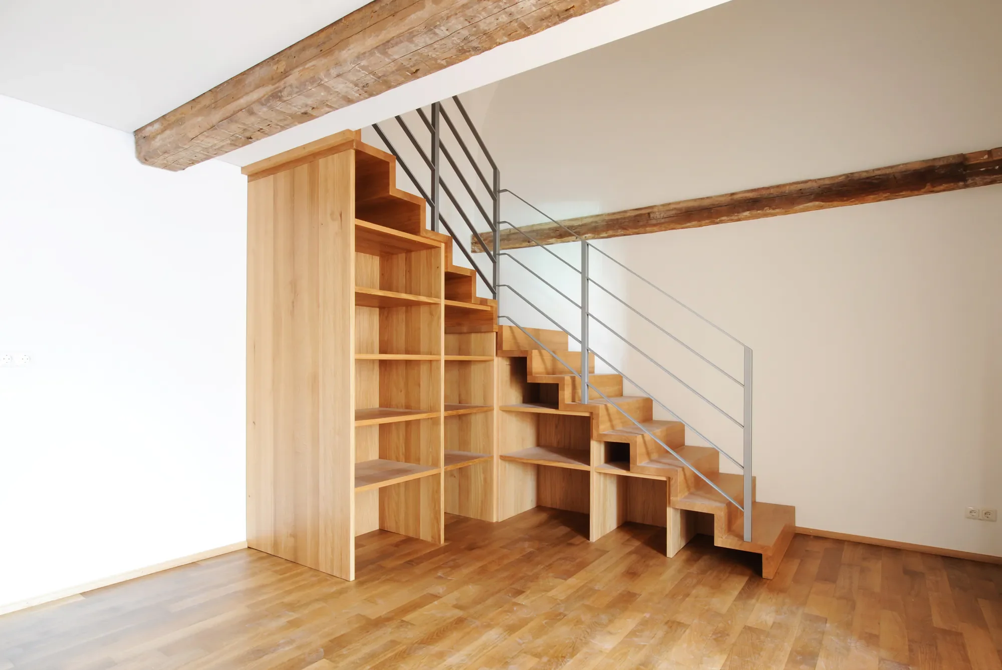 Innenaufnahme eines modernen Treppensystems aus Holz mit Metallhandlauf, integriertem Stauraum unter den Stufen, in einem Raum mit Holzboden und sichtbaren Deckenbalken.