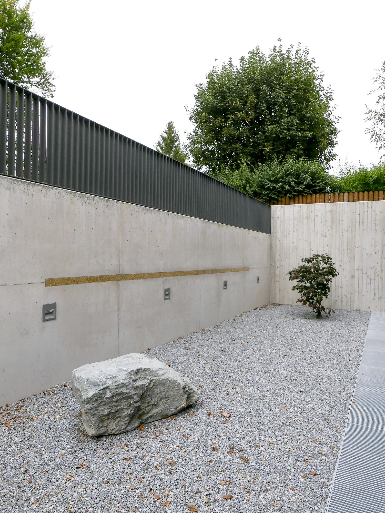 Ein moderner grauer Betonmauer mit dunklem Metallsichtschutz, ein kleiner Baum und ein großer Naturfels im Kiesgarten, im Hintergrund grüne Bäume und ein grauer Himmel.