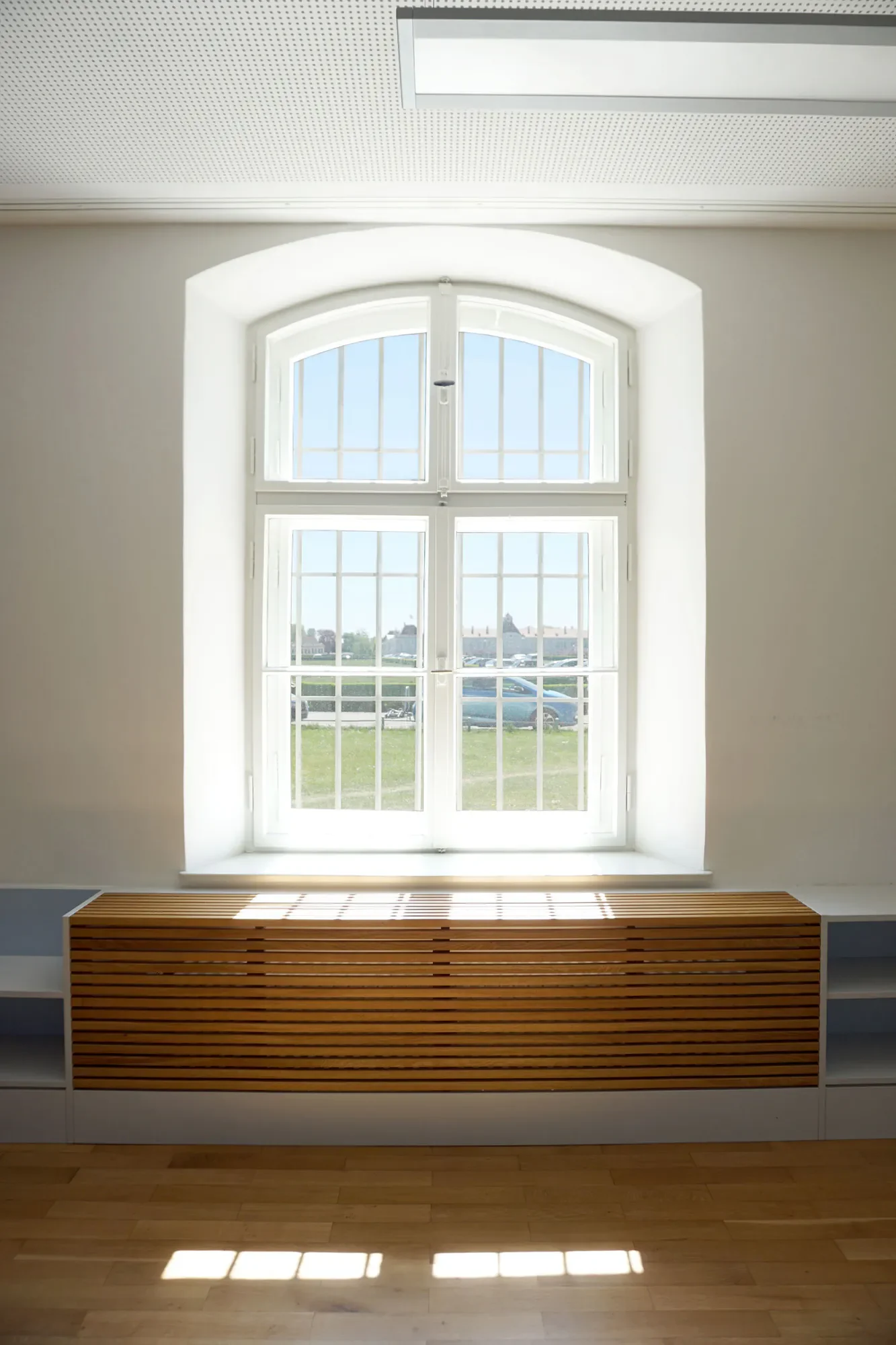 Ein heller Raum mit einem großen, weißen, arched Fenster, durch das das Tageslicht hereinfällt. Unter dem Fenster befindet sich eine gepolsterte Sitzbank aus Holzlamellen. Im Hintergrund sind grüne Wiesen und Gebäude sichtbar.