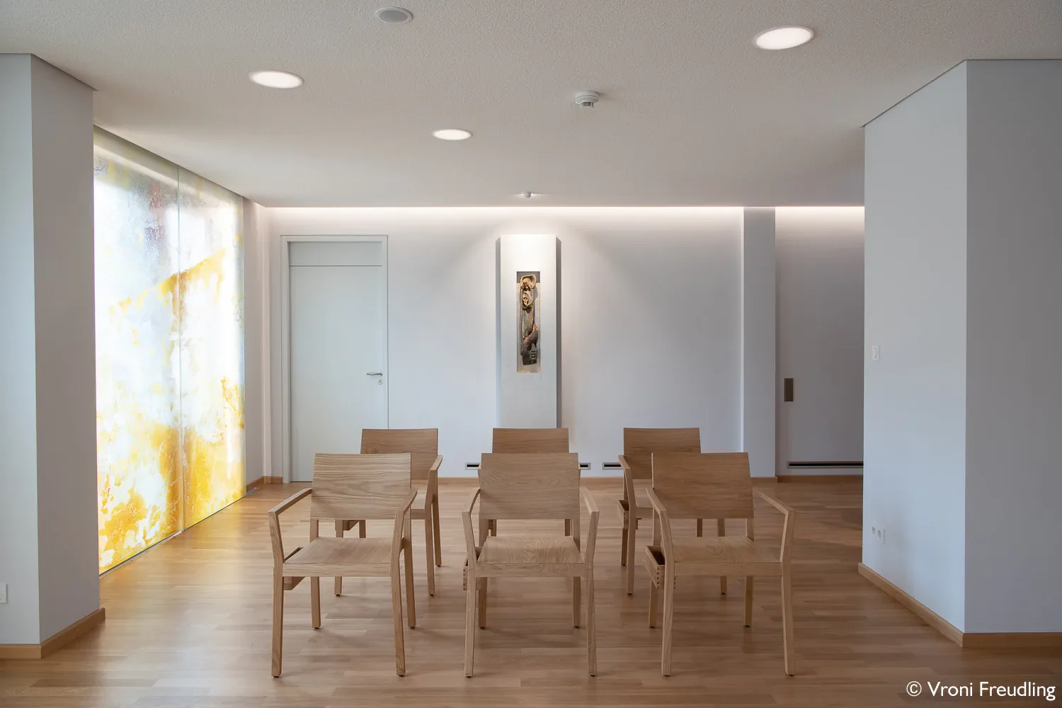 Ein Raum mit weißen Wänden und Holzboden, vier Holzstühle in einer Reihe, eine weiße Tür, eine vertikale Kunstinstallation an der Wand, eine beleuchtete Decke, eine große gelbe-blumige Verpackung oder Vorhang.