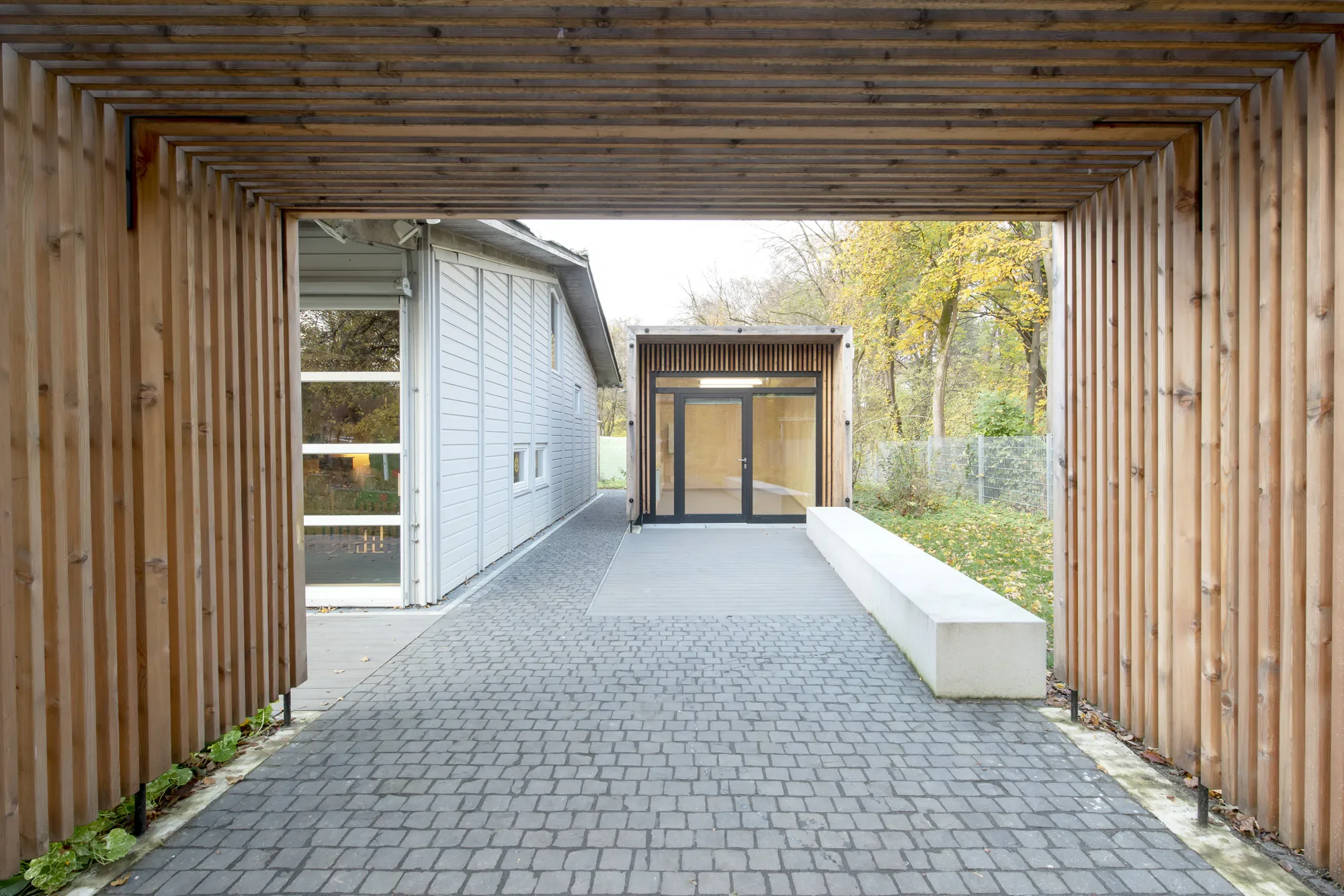 Blick durch einen holzverkleideten Eingangsbereich auf einen gepflasterten Weg zu einem modernen Gebäude mit Glas und Holz, umgeben von Laubbäumen.