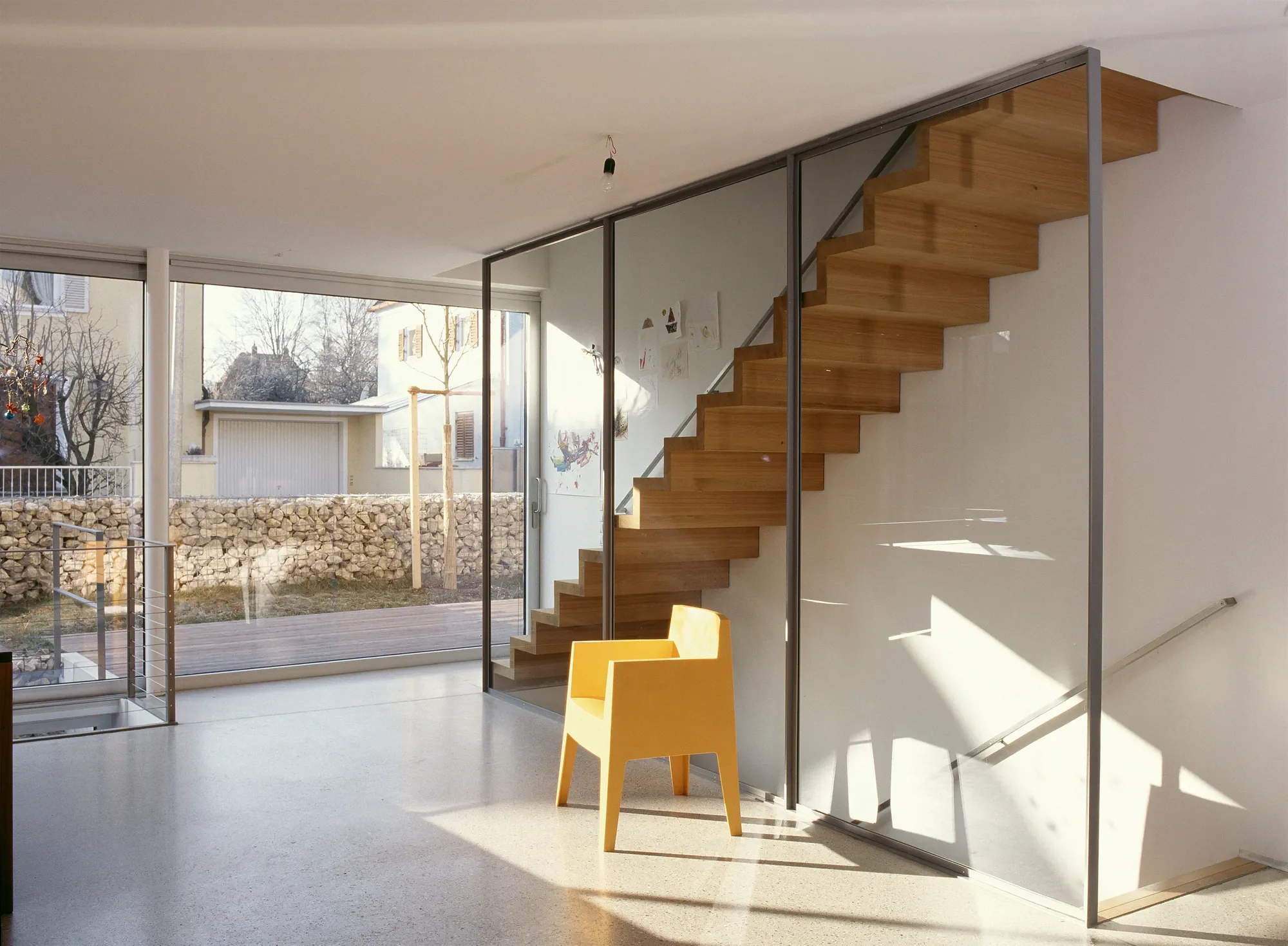 Innenraum eines modernen Hauses mit offener Treppe aus Holz und Glas, großer Fensterfront mit Blick auf den Garten, minimalistische Einrichtung, gelbe Stühle, natürliche Lichtverhältnisse.