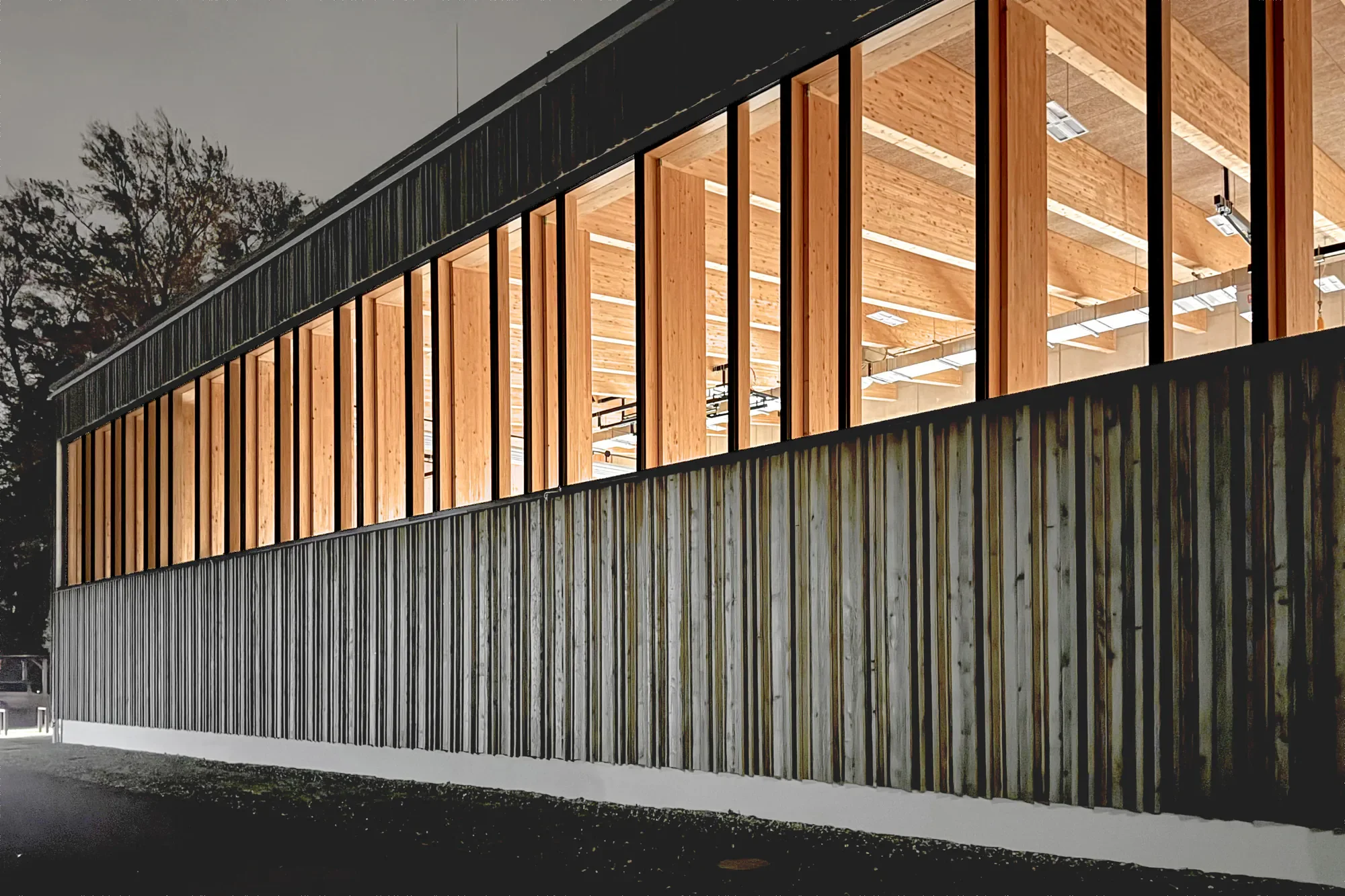 Moderne Gebäudefassade mit vertikal angebrachten Holzlamellen und großen Fenstern, beleuchtet bei Nacht.