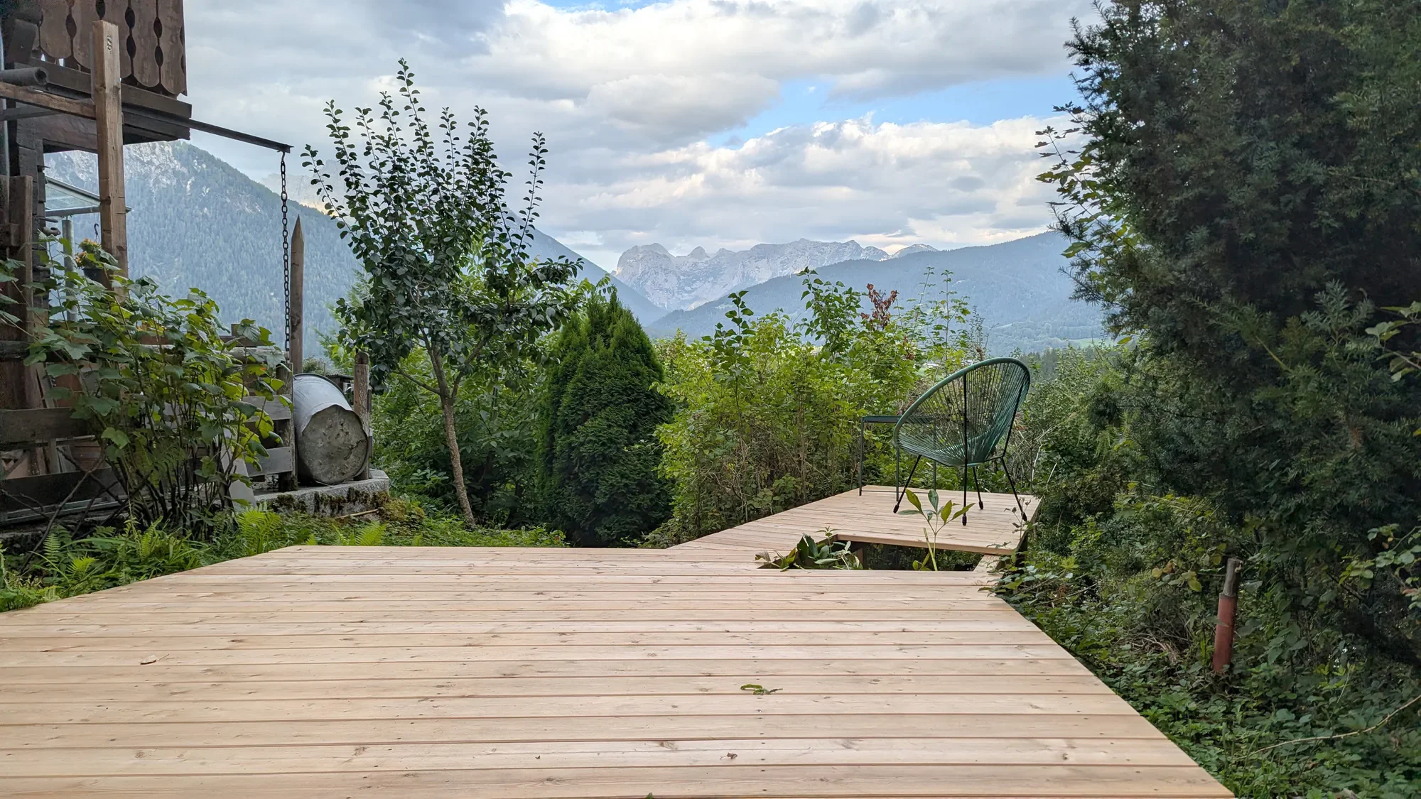Ein Holzdeck mit einem schwarzen Stuhl, umgeben von Bäumen und Pflanzen, mit Blick auf Berge im Hintergrund.