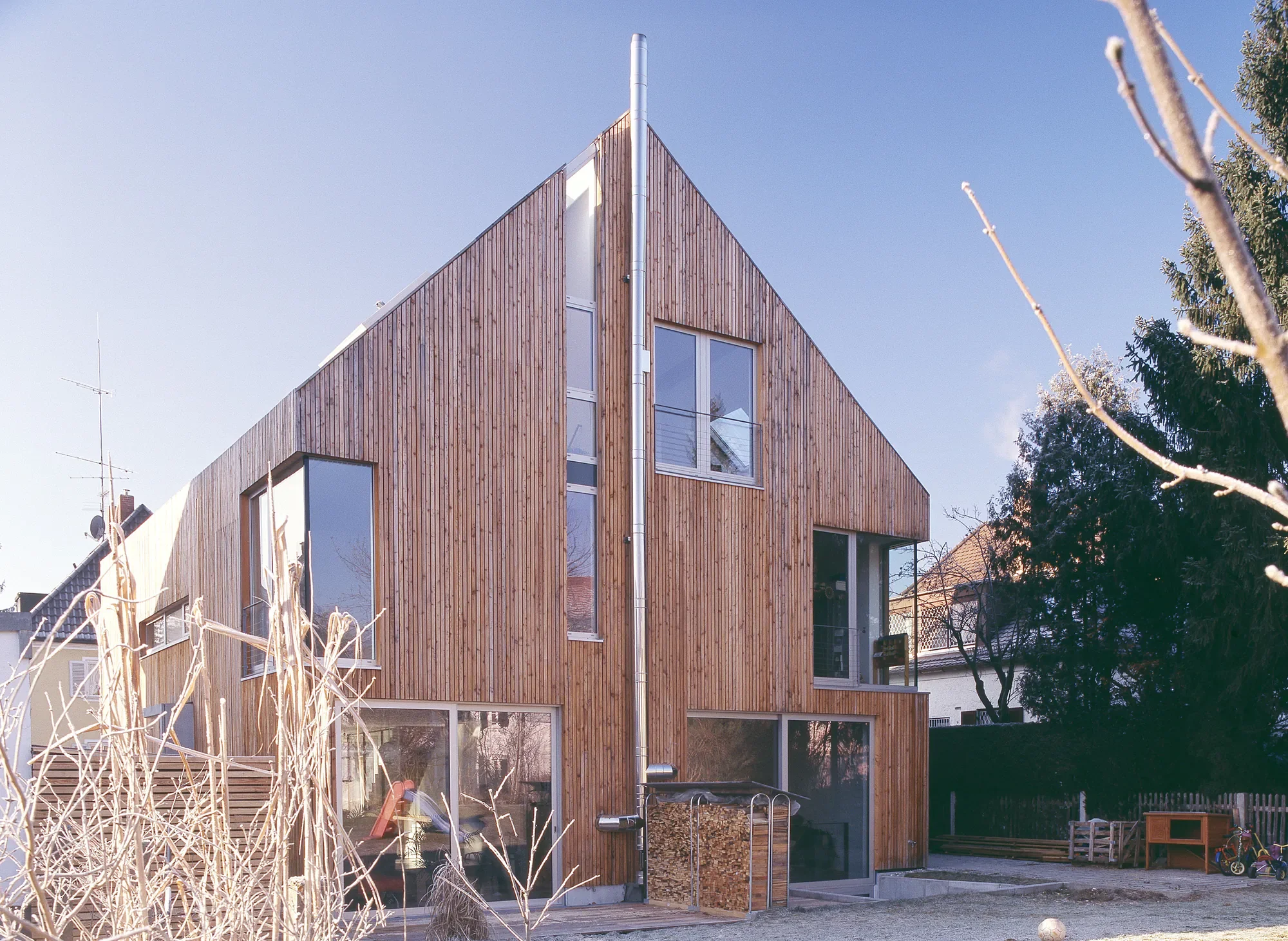 Moderne gebaute holzfassade Mehrfamilienhaus mit mehreren Etagen, großen Fenstern und einem Sichtschacht für die Abwasserleitung, umgeben von einem Garten mit Bäumen und Spielzeug.