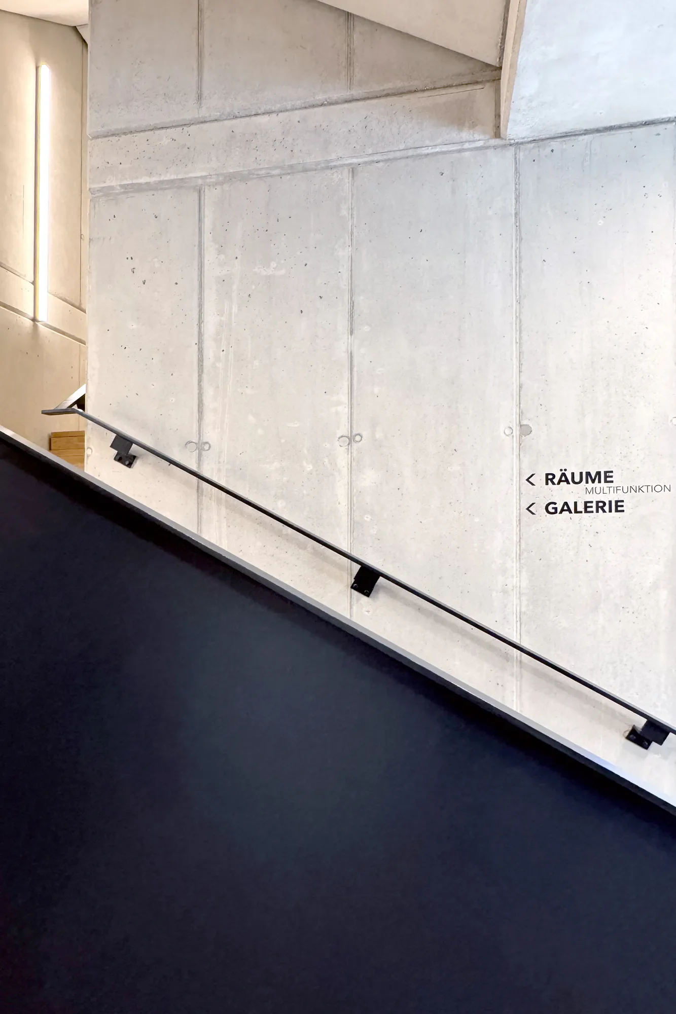 Ein Blick auf eine Treppe mit schwarzem Teppich und einer schwarzen Handläufe, neben einer weißen Betonwand mit Beschriftungen, die auf Räume und eine Galerie hinweisen.