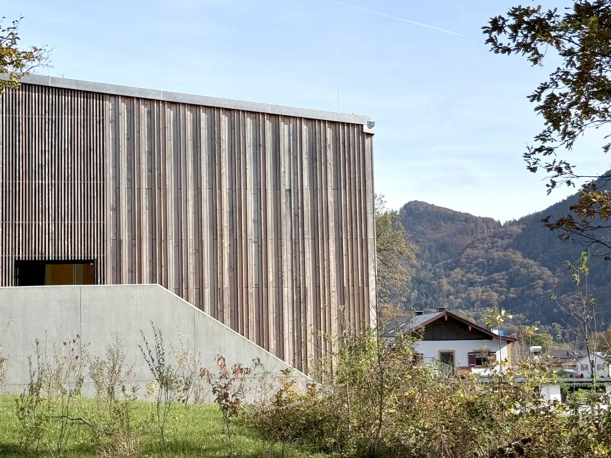 Ein modernes Gebäude mit vertikalen Holzverkleidungen, umgeben von Bäumen und Bergen im Hintergrund bei sonnigem Wetter.