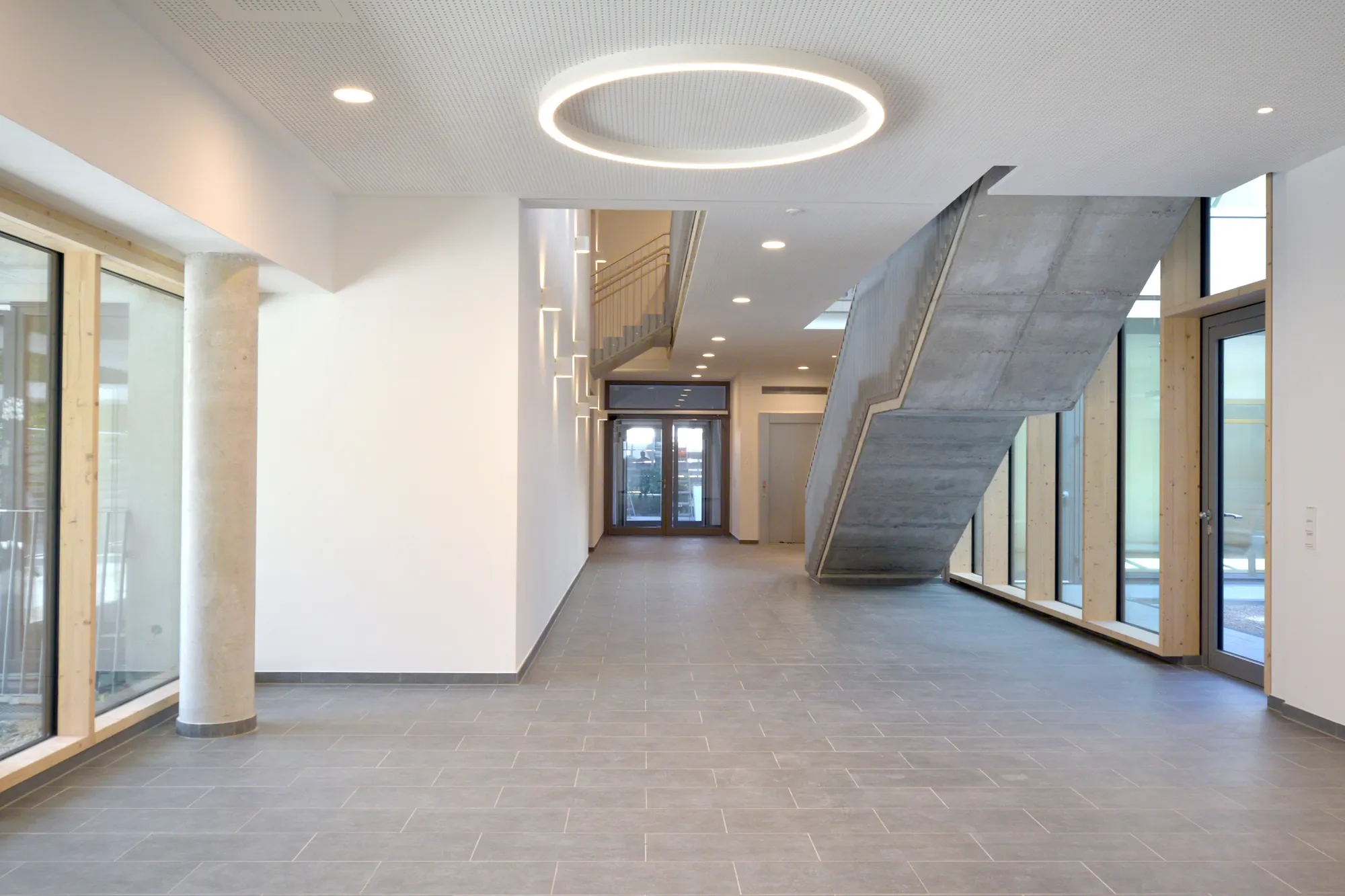 Innenansicht eines modernen Gebäudes mit einem Treppenaufgang aus Beton, großen Fenstern und hellem Licht.