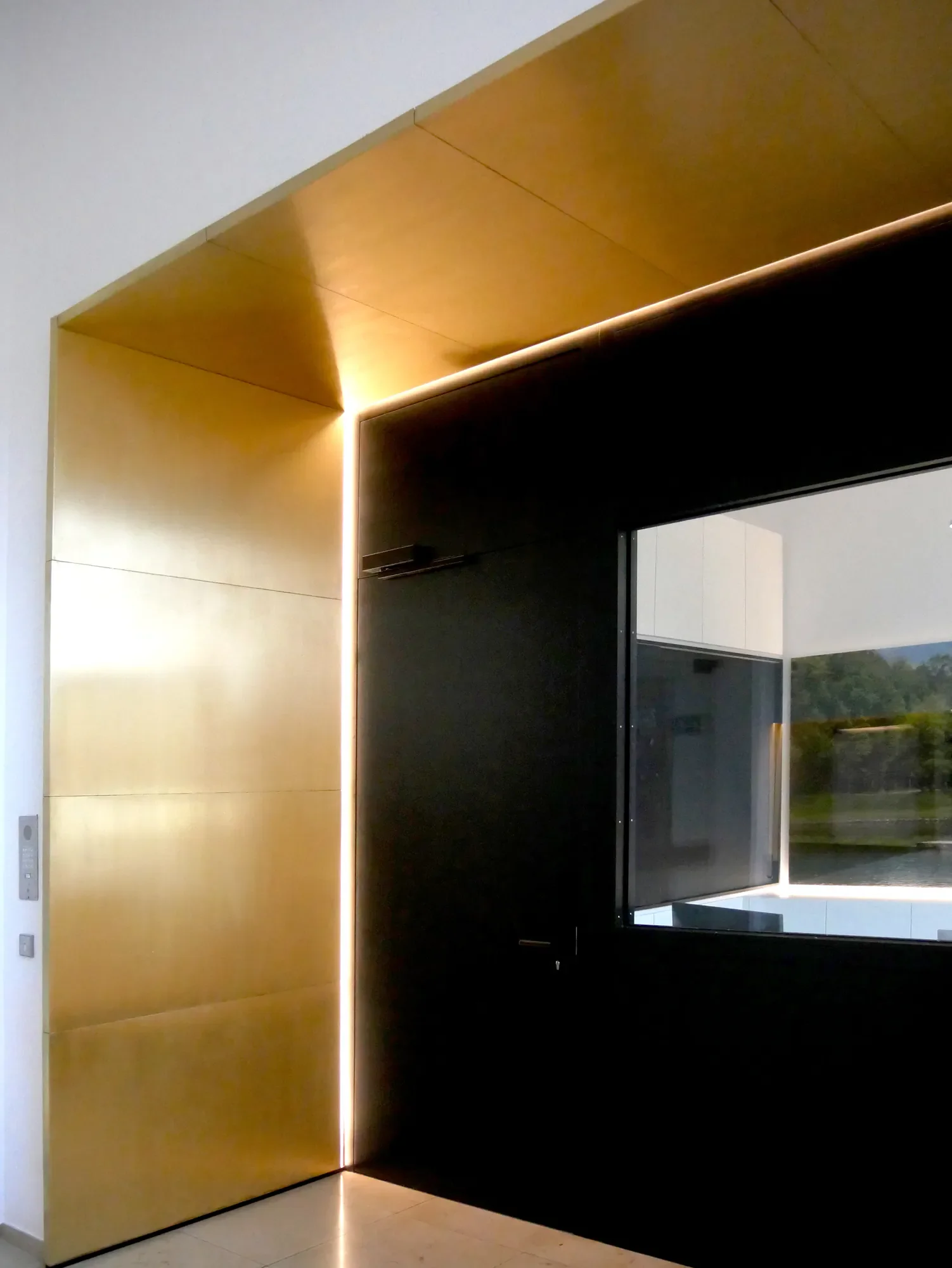 Ein moderner Innenraum mit goldener Wand, schwarzem Wandabschnitt, einem großen Fenster und einer Sicht nach draußen, mit natürlichem Licht und Landschaft im Hintergrund.